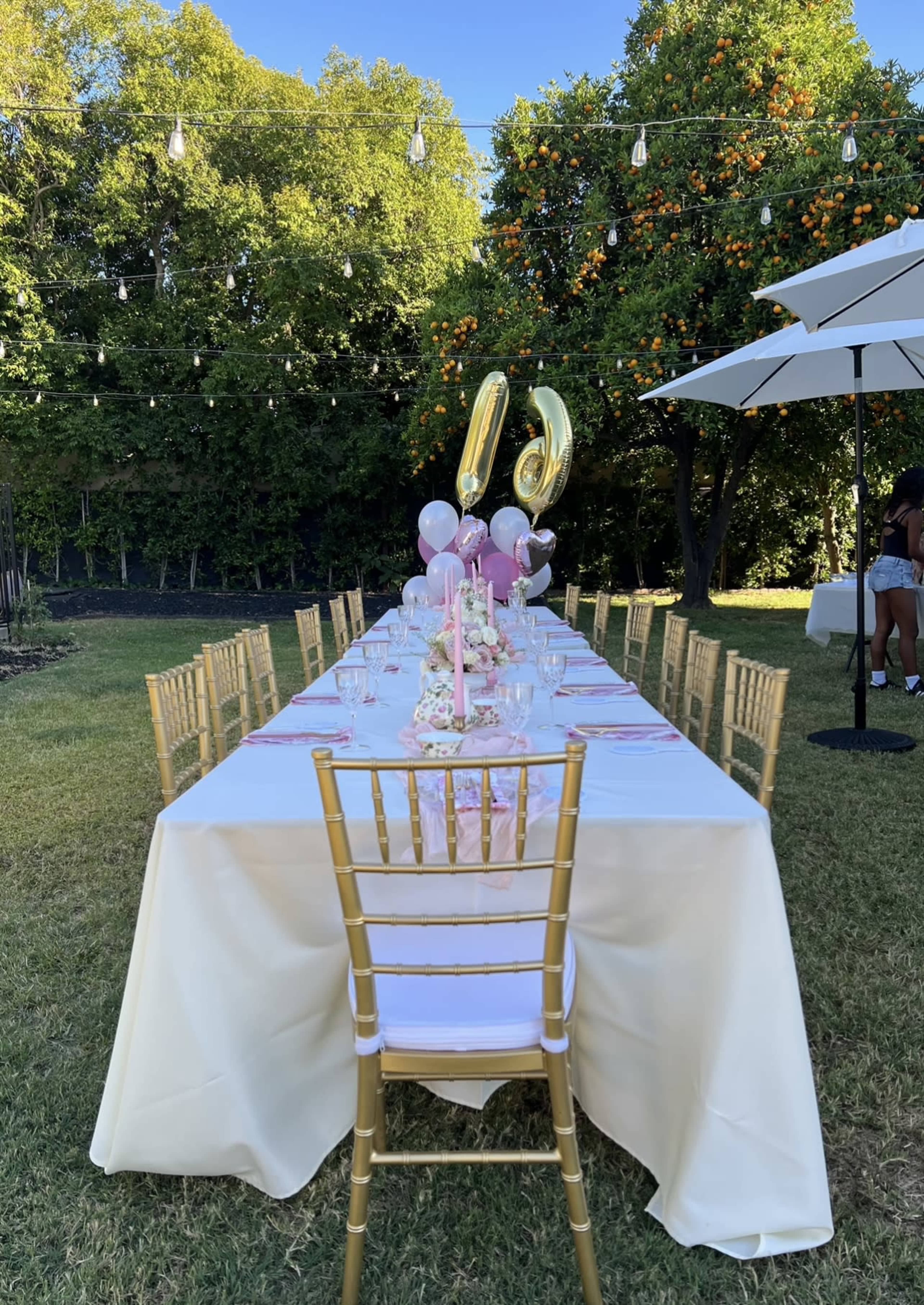 A long dining table is set outdoors with pink decorations, golden balloons, and chairs under string lights and orange trees.