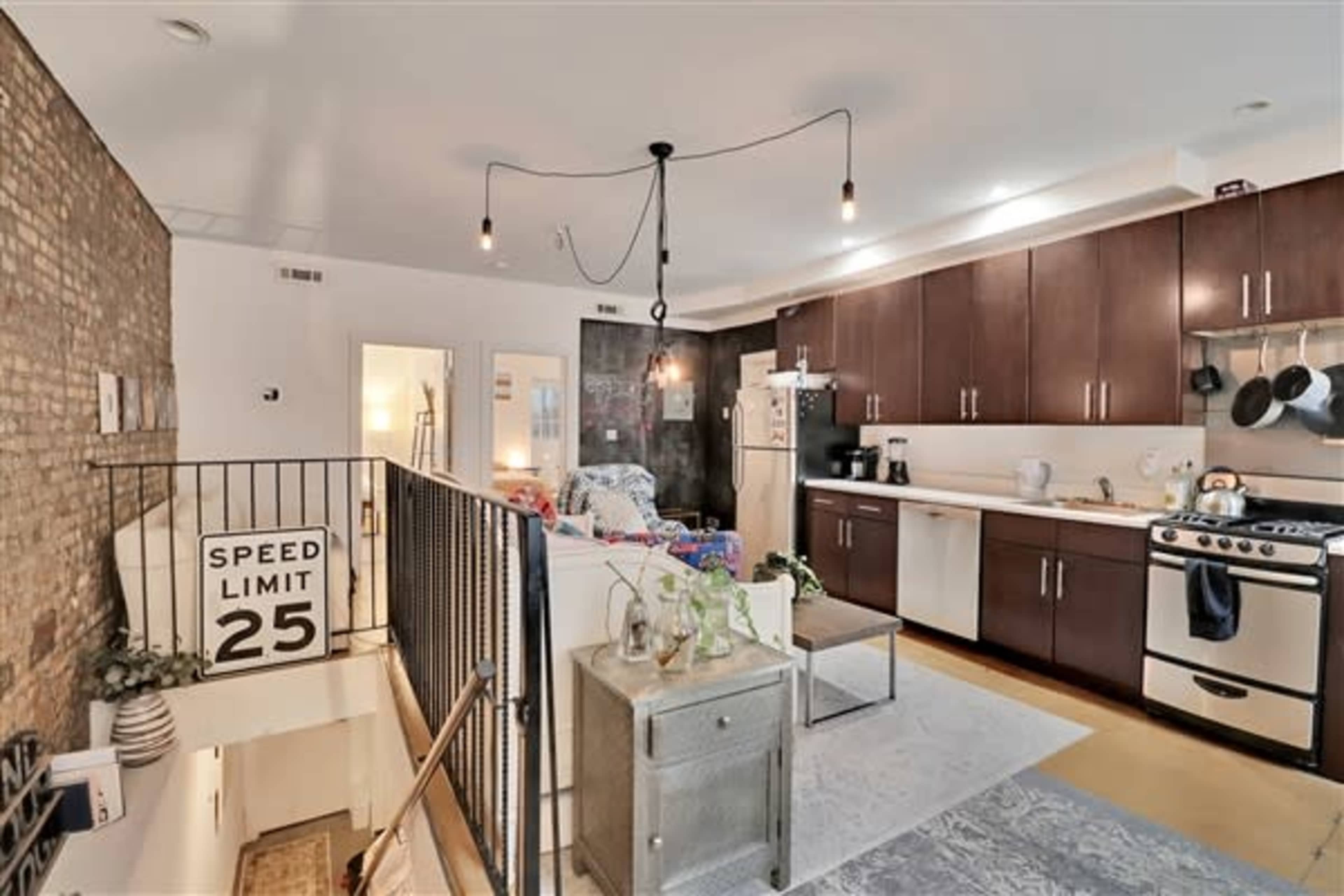 A modern kitchen with dark wooden cabinetry, a white refrigerator, and a stove, adjacent to a living area featuring exposed brick walls and a staircase.
