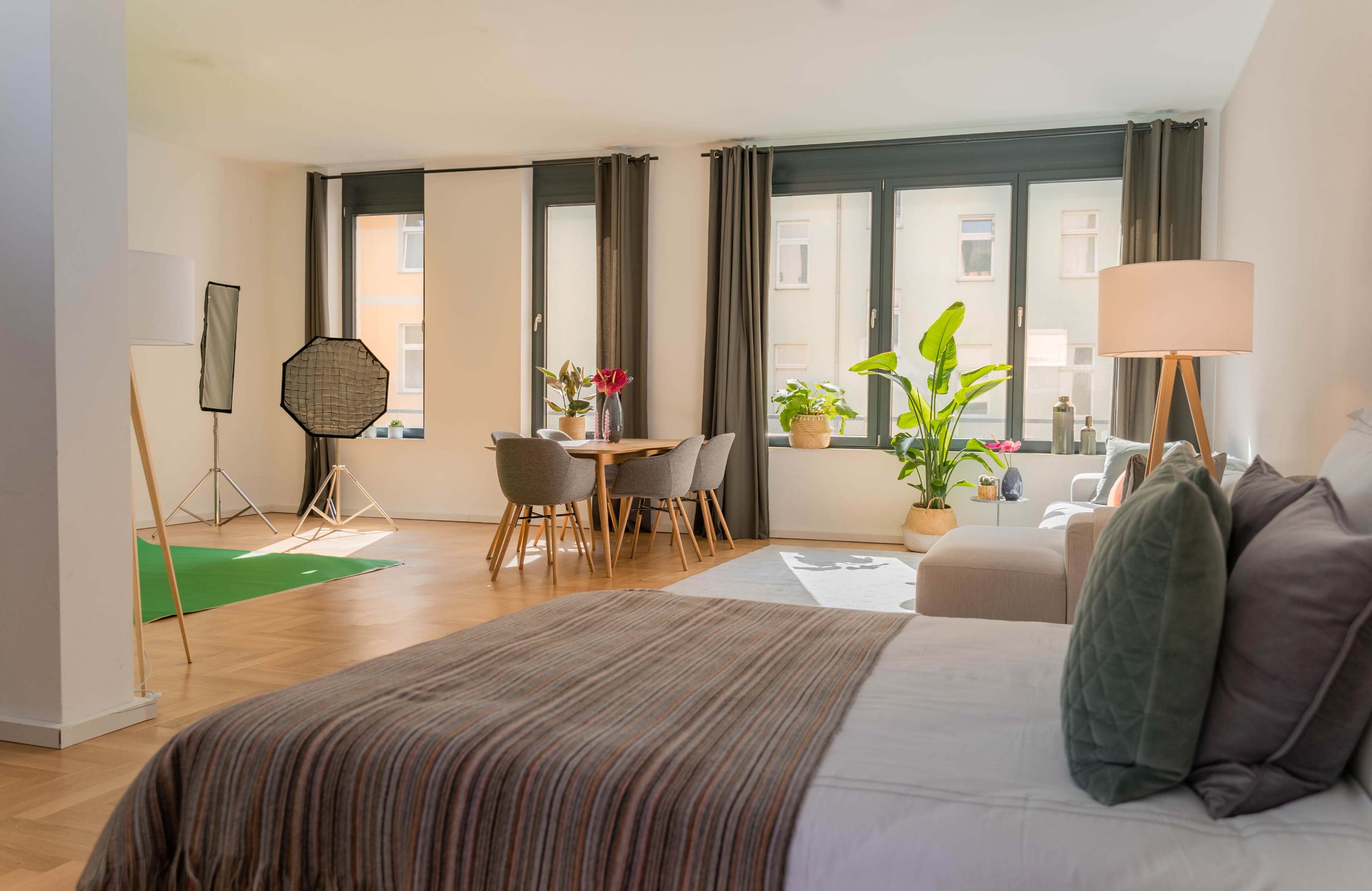 A spacious, well-lit apartment featuring a seating area with a sofa, a dining table with chairs, and large windows with plants.