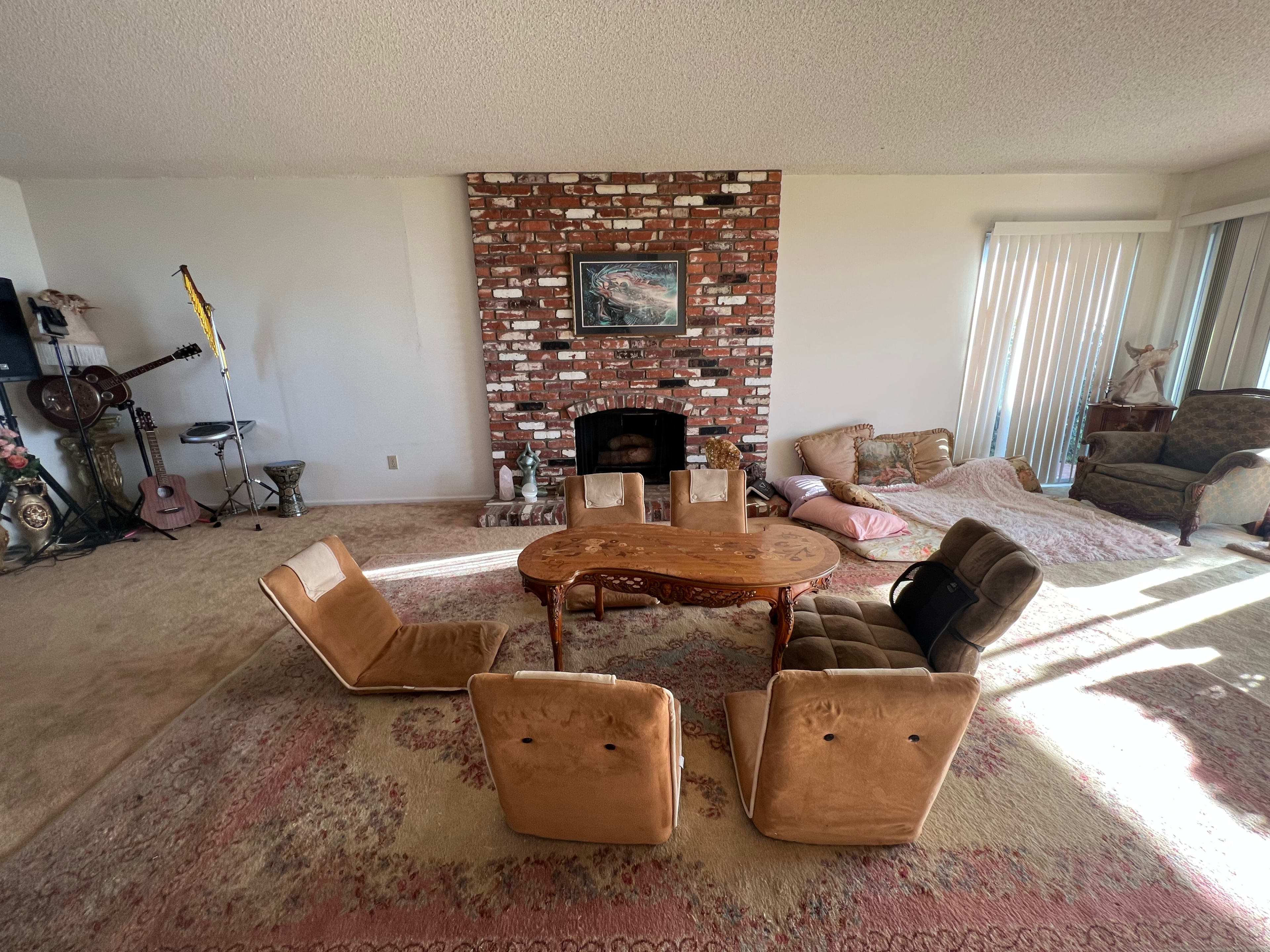 A spacious living room features a circular wooden table surrounded by four beige reclining chairs, with a brick fireplace and various seating arrangements in the background.
