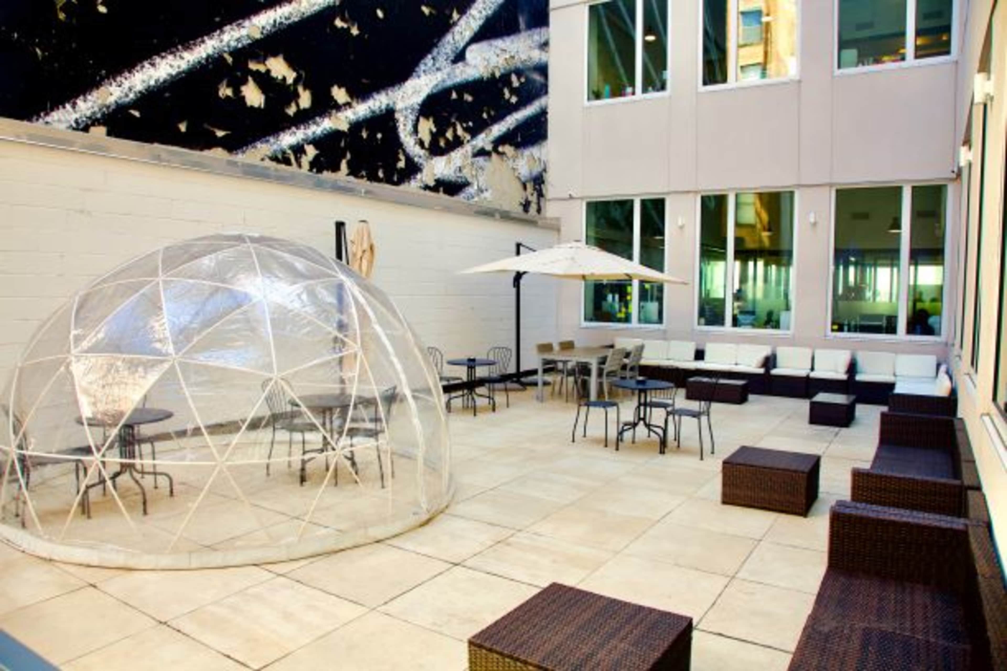 A courtyard with a transparent dome structure, several tables and chairs, and a large umbrella for shade.