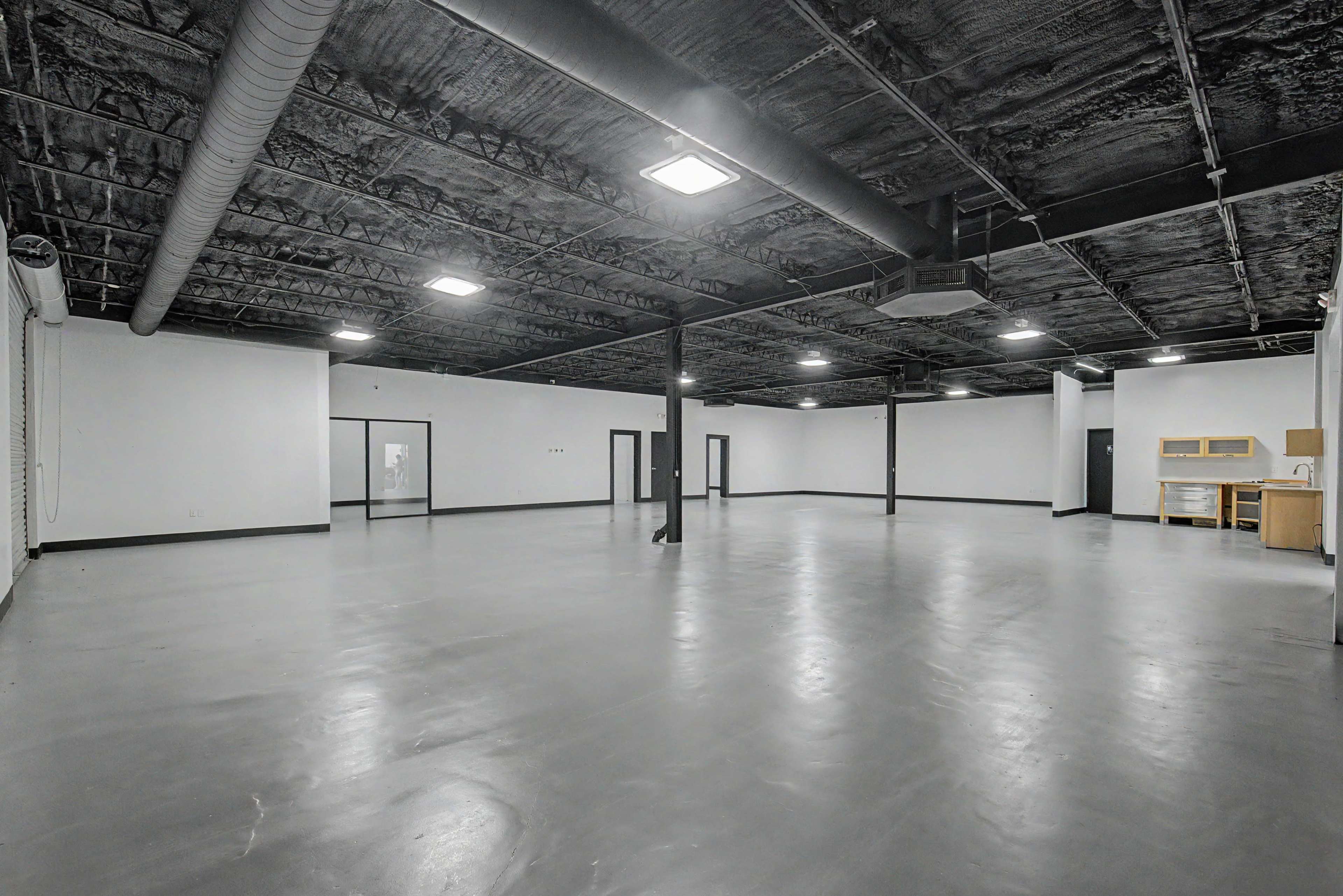 The image shows a large, empty commercial space with a gray concrete floor and exposed black ceilings, featuring several doorways and a kitchenette area in the background.