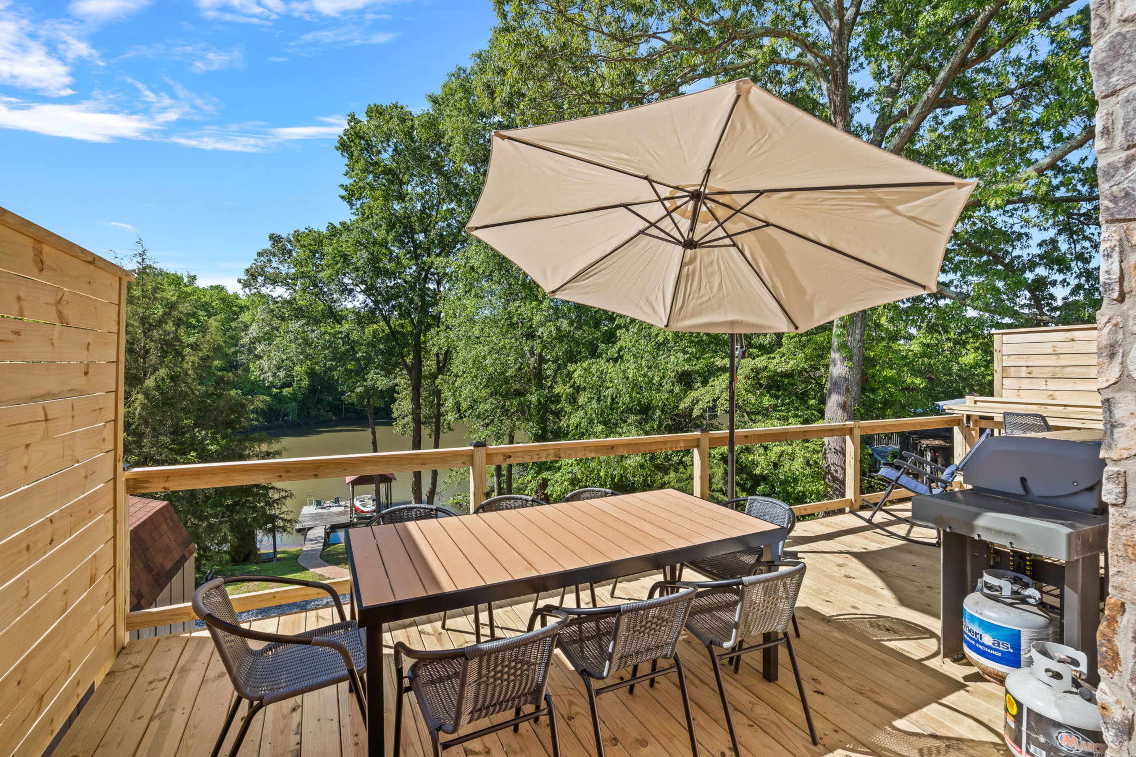 A wooden deck with a table and chairs is shaded by a large umbrella, overlooking a calm body of water surrounded by trees.