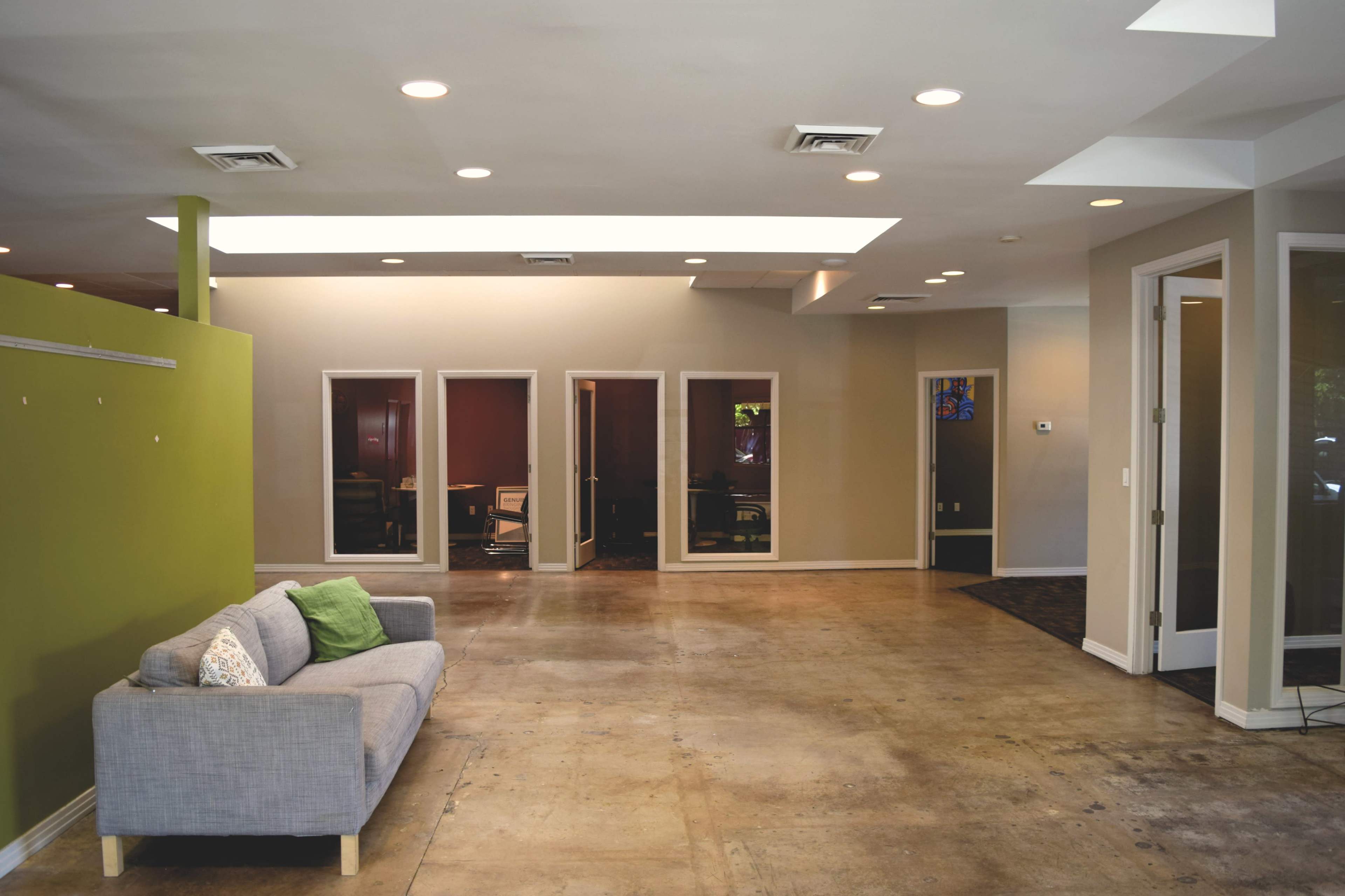 The image shows a spacious, modern office interior with a gray sofa, green accent wall, and several glass-enclosed meeting rooms visible in the background.