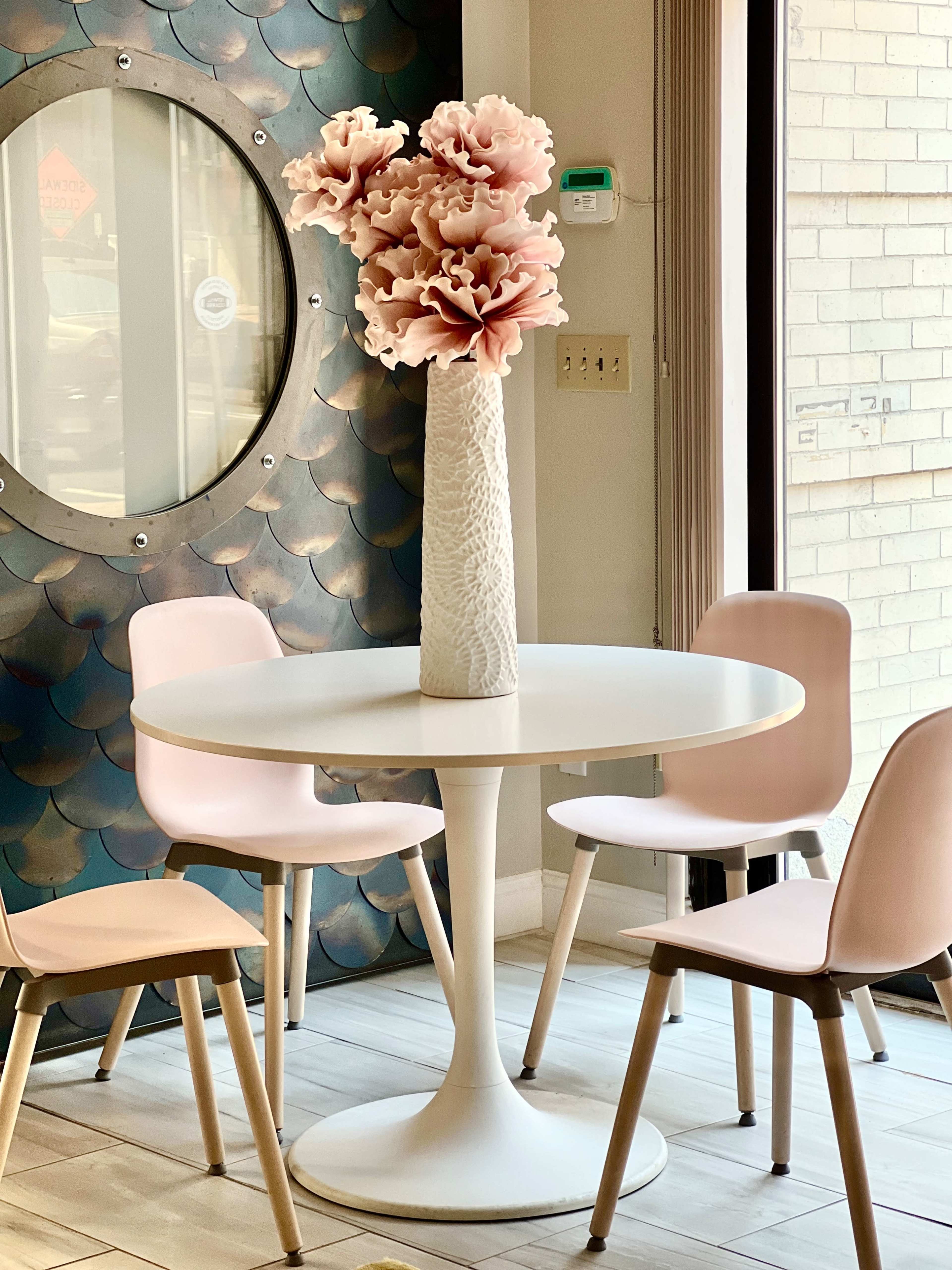 A round white table with four chairs sits near a wall decorated with a circular mirror and a textured design, featuring a large vase filled with pink flowers at its center.
