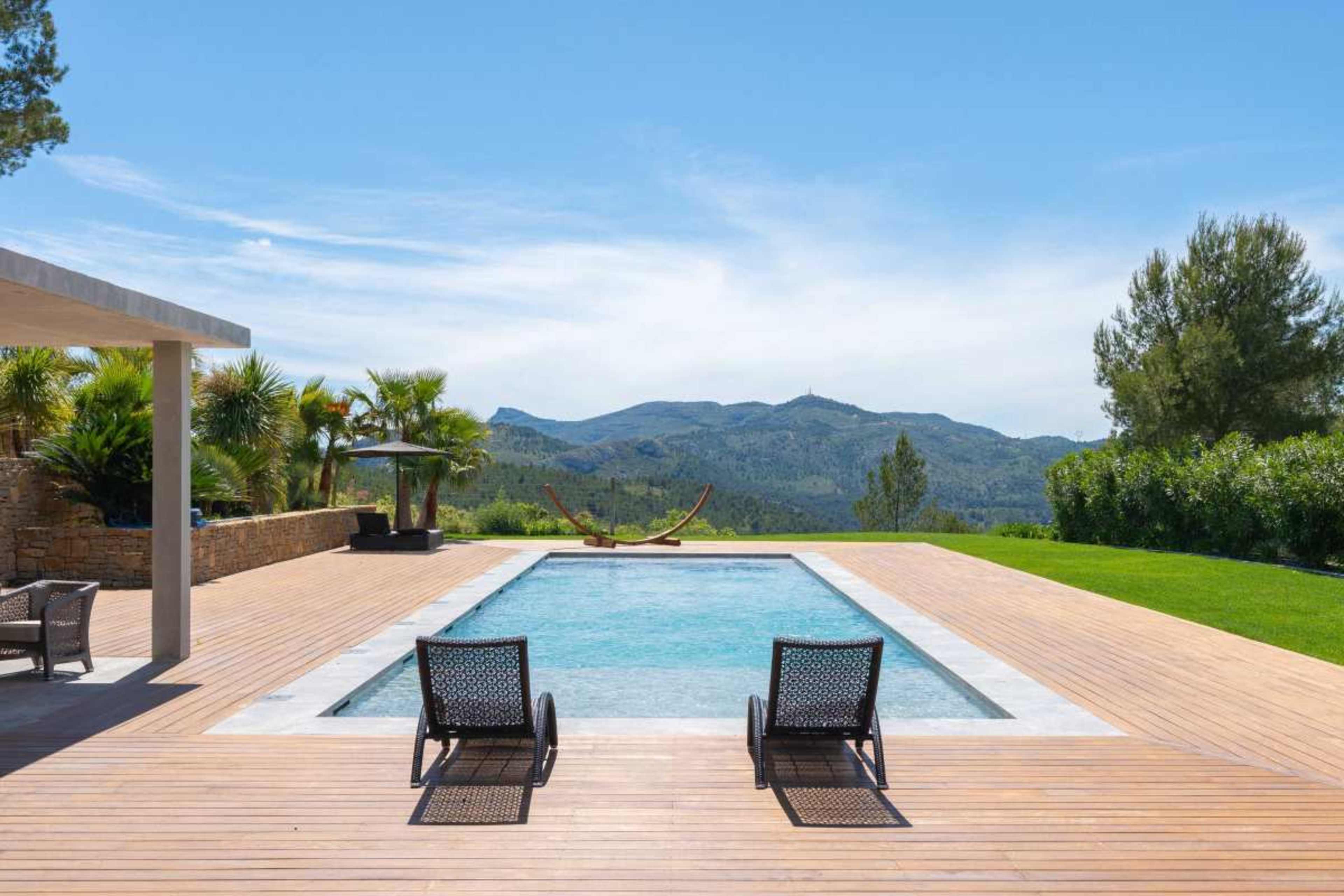 A rectangular swimming pool is situated on a wooden deck with two lounge chairs, surrounded by greenery and a view of distant mountains under a clear blue sky.