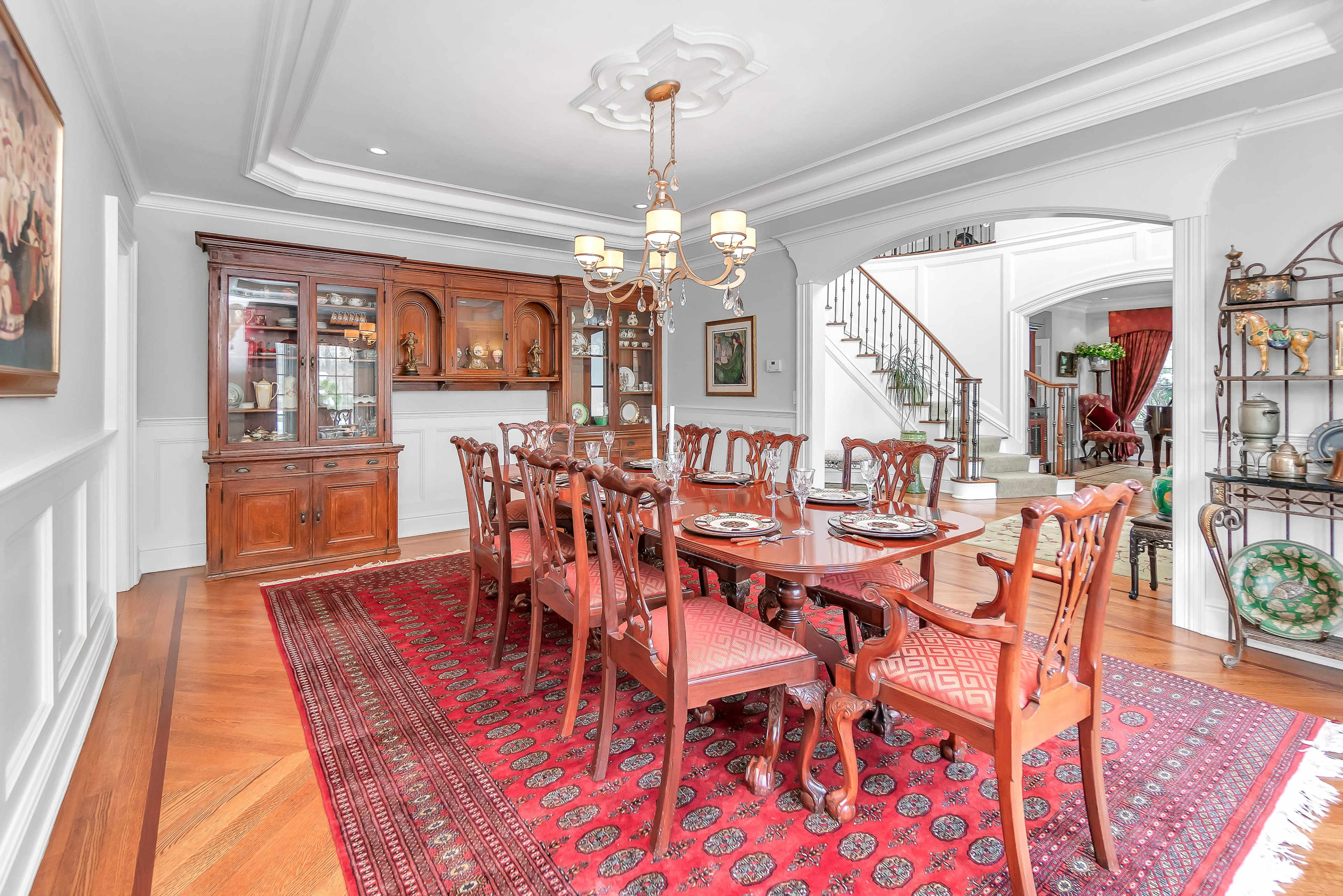 A formal dining room with a large wooden dining table surrounded by chairs, a china cabinet, and a decorative rug on the hardwood floor.