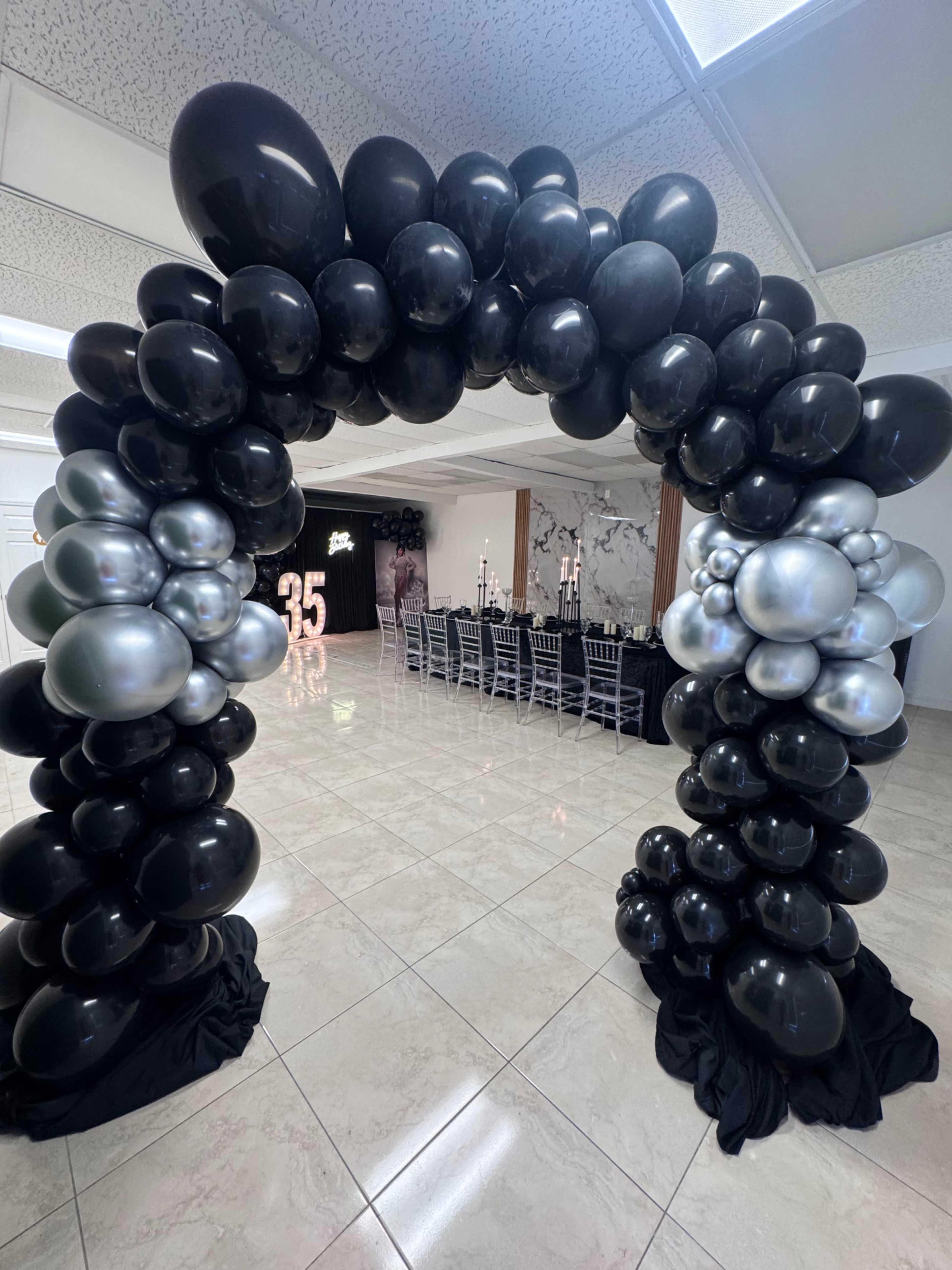 A black and silver balloon arch stands at the entrance of a decorated event space featuring a long table set for a celebration.