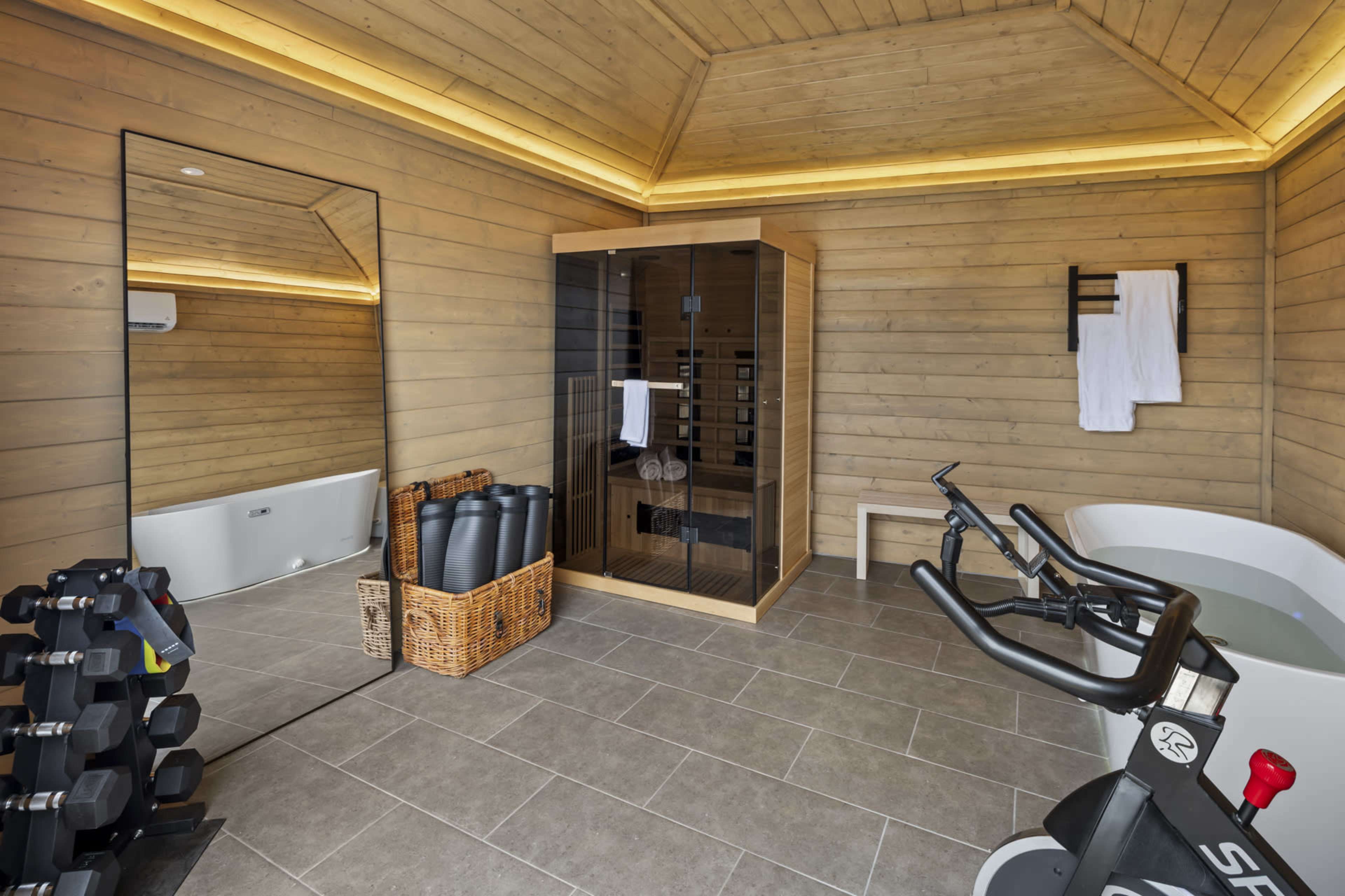 The image shows a wooden interior spa area featuring a sauna, a stationary bike, a basket of exercise mats, a mirror, and a freestanding bathtub.