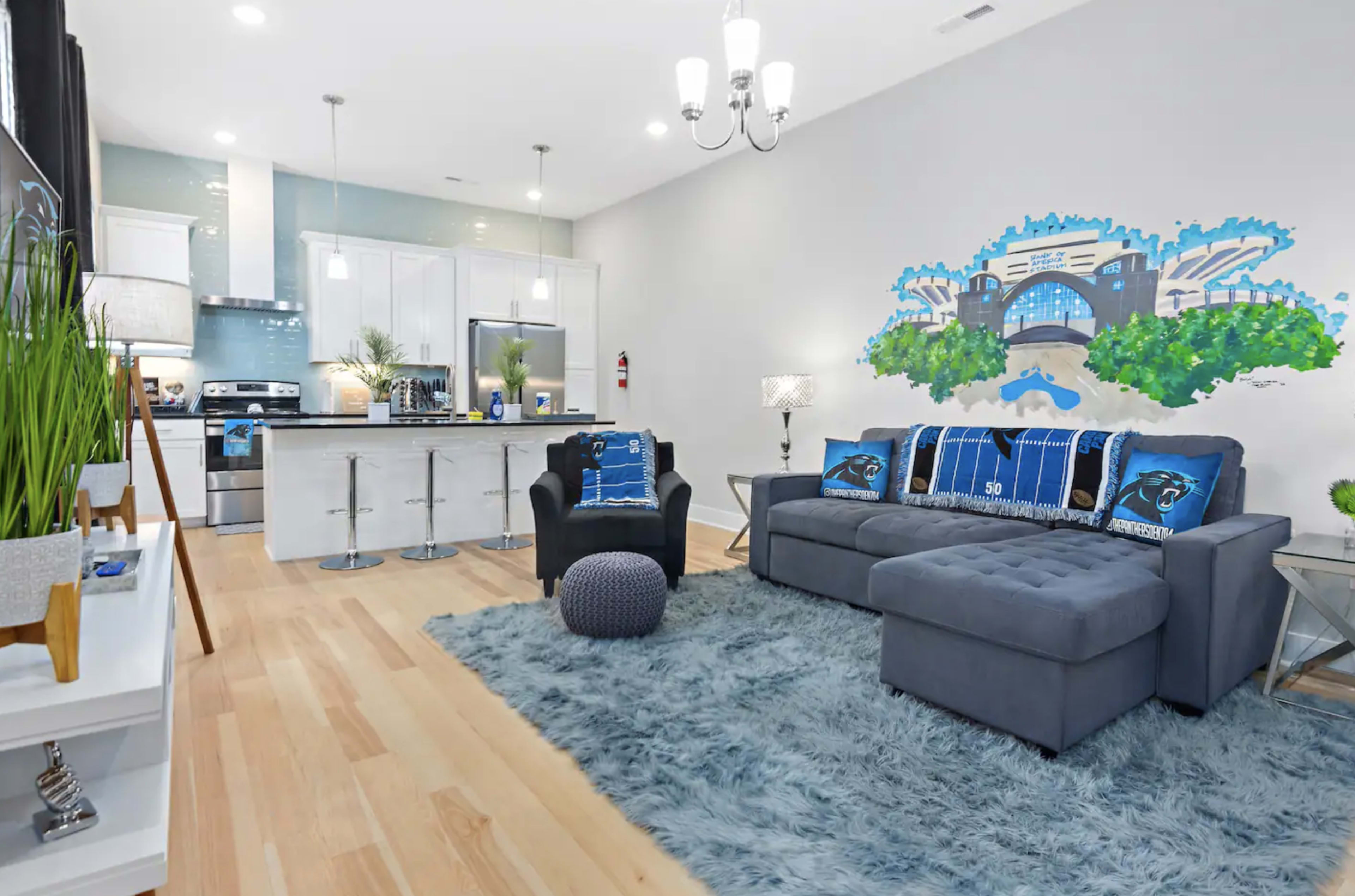 A modern living area featuring a gray sofa, a light blue rug, and a kitchen with white cabinetry, along with a mural of a cityscape on the wall.