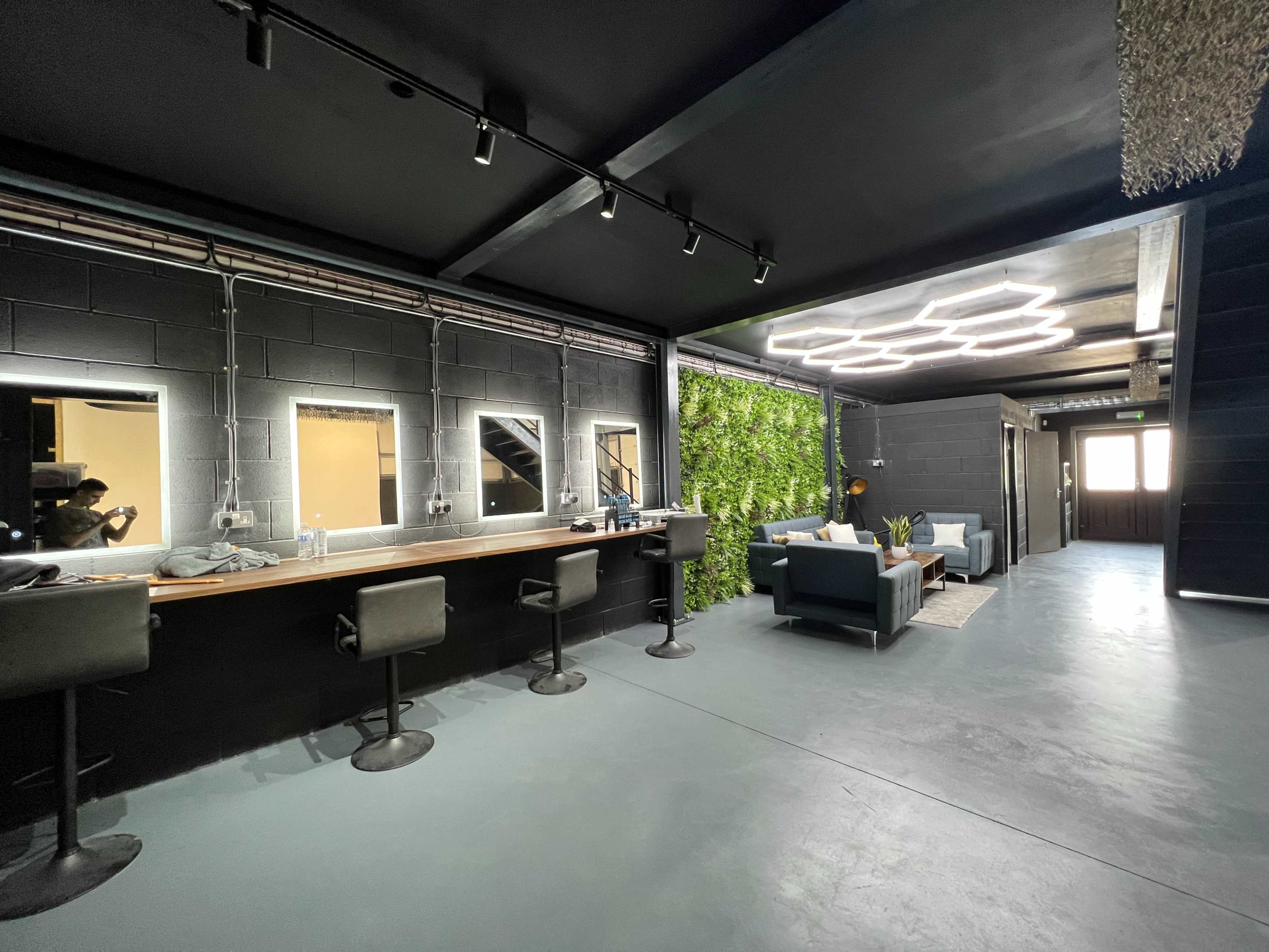 The image shows a modern interior space with a sleek seating area, a long counter with mirrors, and a green wall feature.