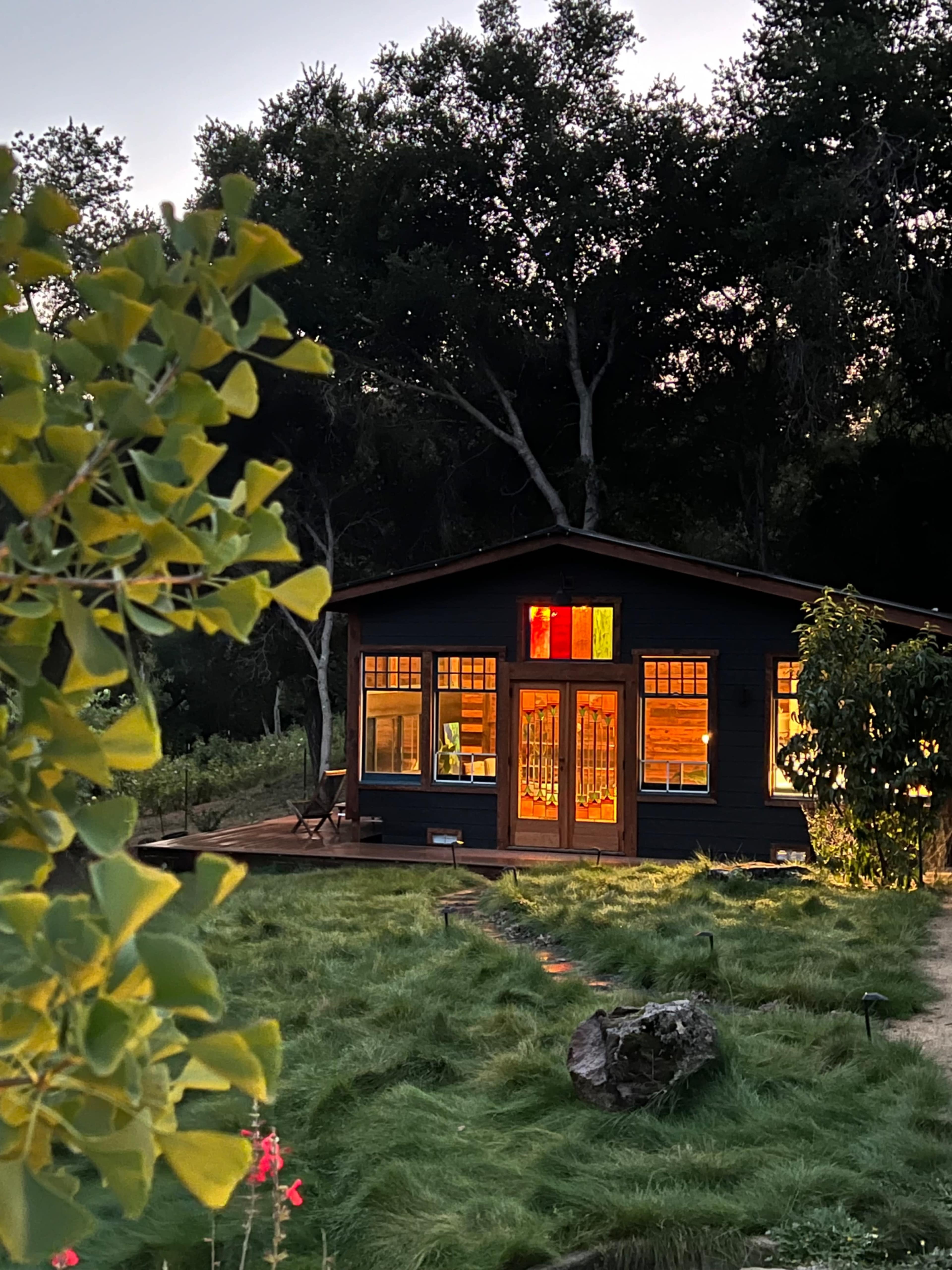A dark-colored house with bright windows is surrounded by lush grass and trees during twilight.