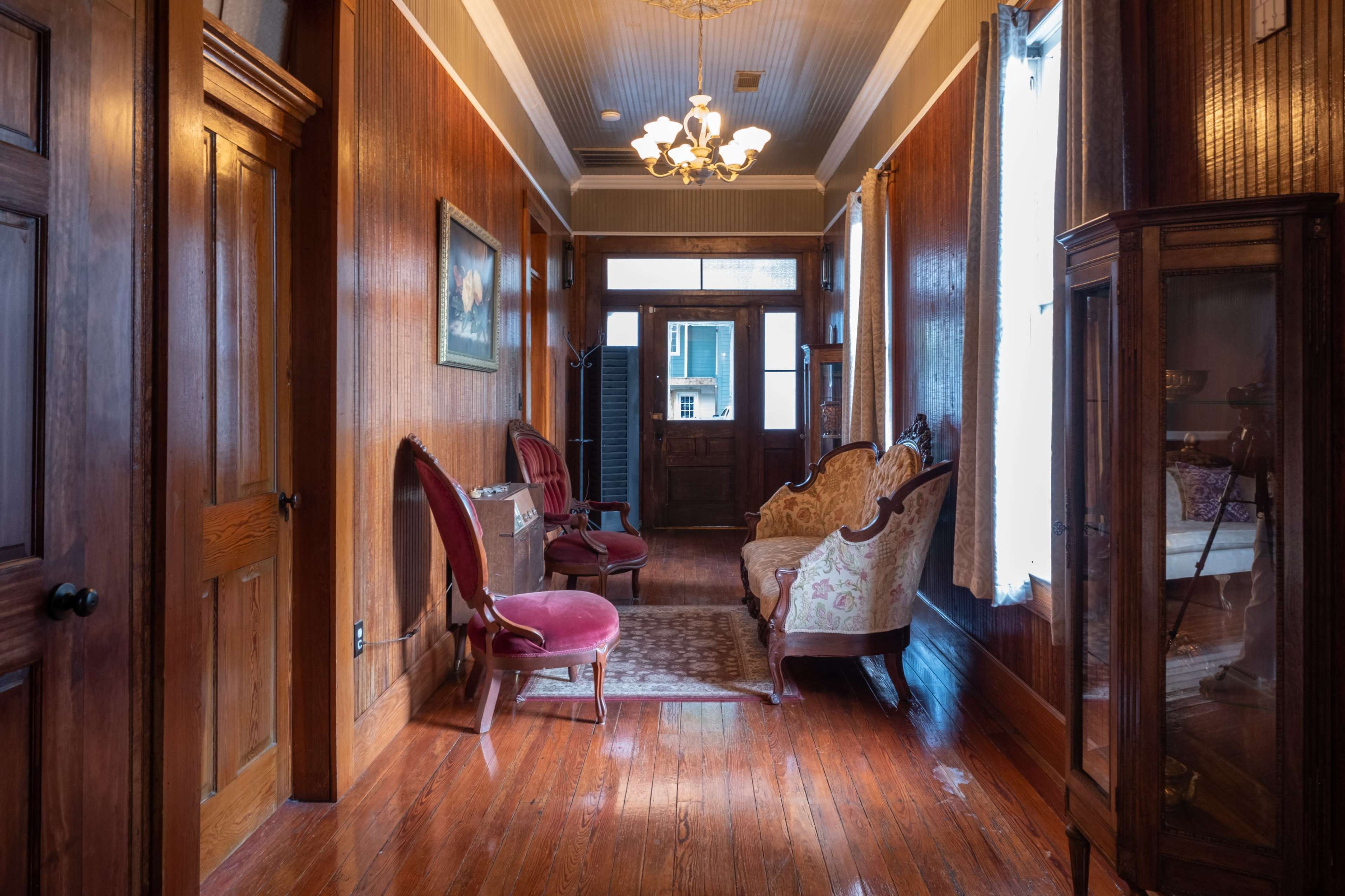 The image shows a spacious hallway featuring wooden walls, a patterned rug, vintage seating, and a chandelier, with a door leading outside.