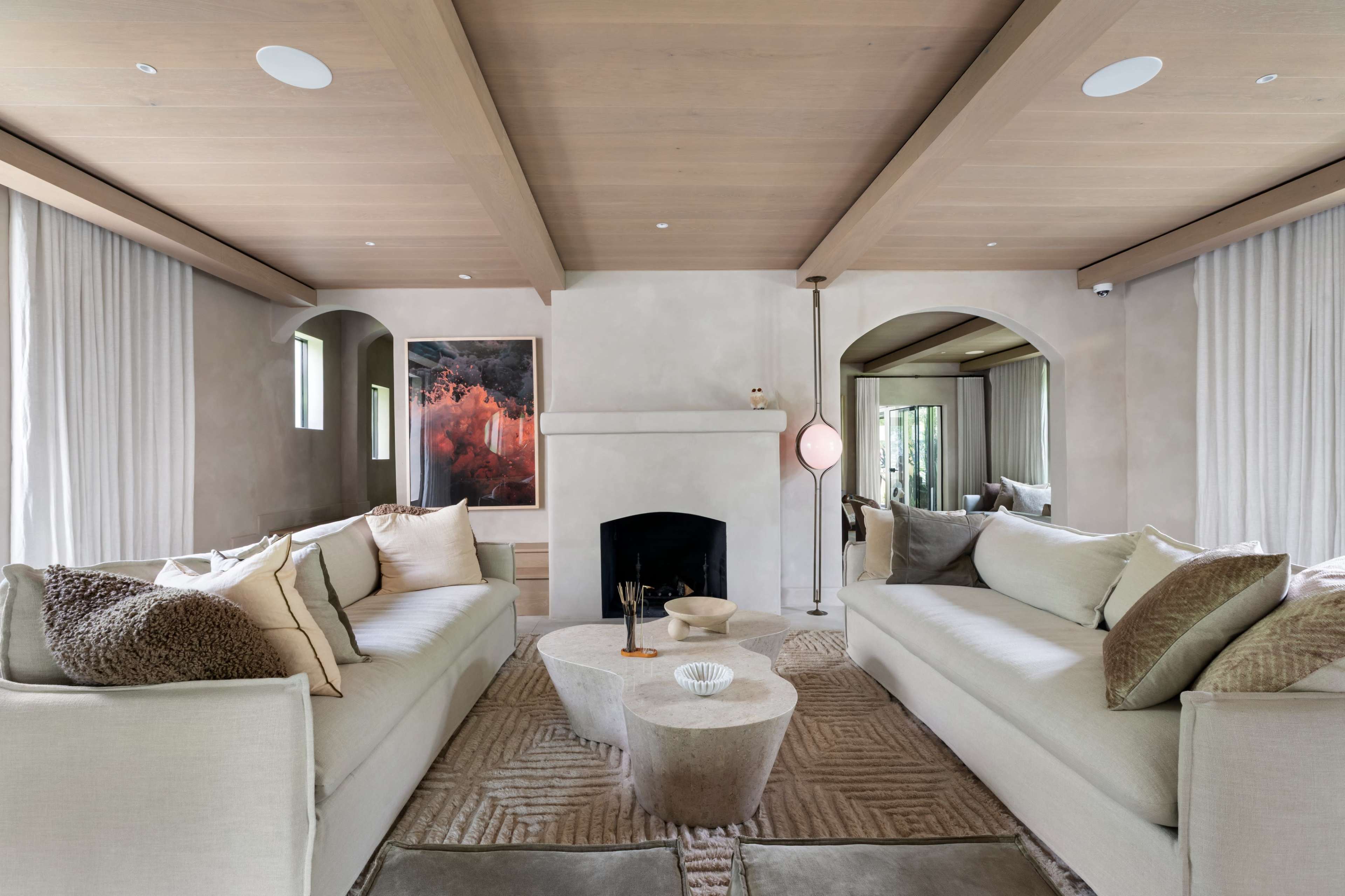 The image shows a modern living room featuring two sofas facing each other, a central round coffee table, and a fireplace framed by light-colored walls and wooden beams.