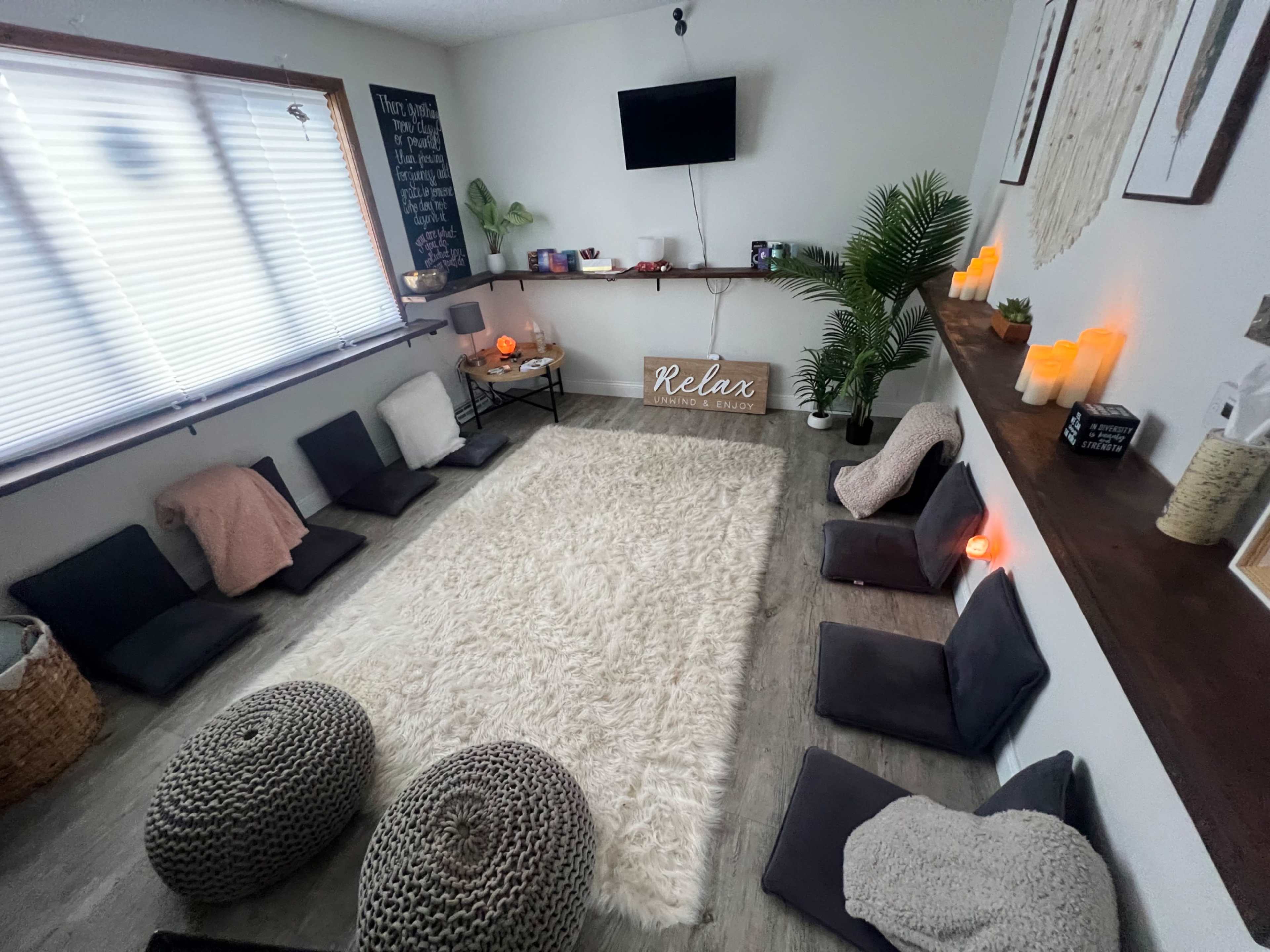 A cozy, minimalist relaxation room features floor cushions arranged around a large, soft rug, with candles and a TV on the wall.