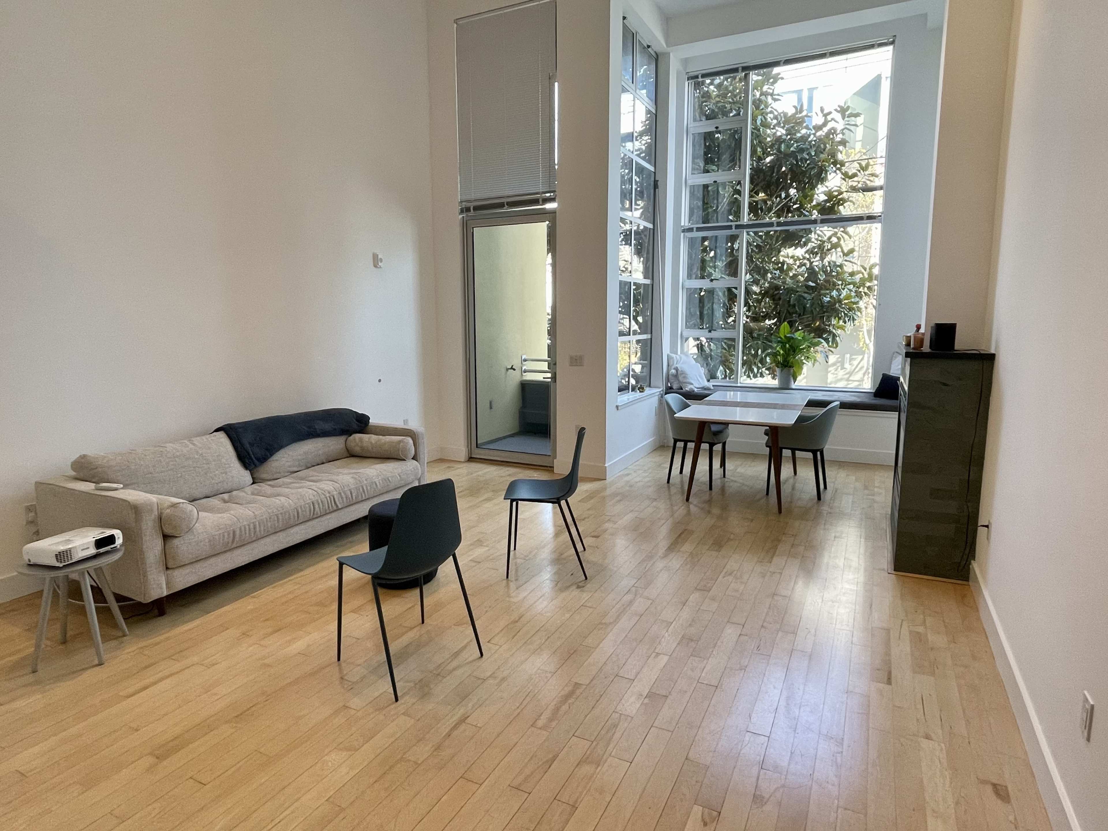 A spacious room with a beige sofa, a small table with four chairs, and large windows overlooking a garden.