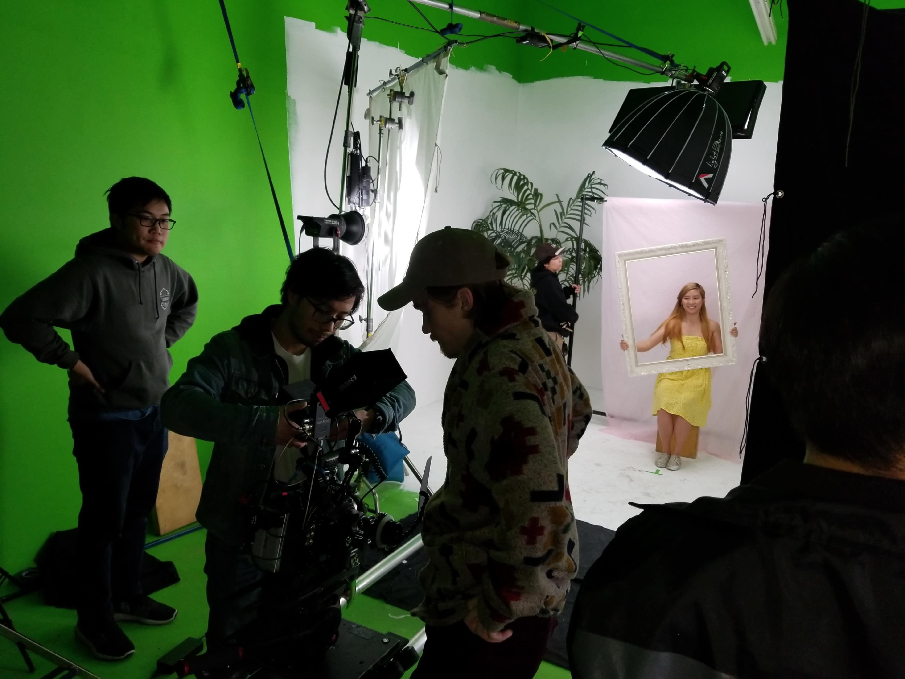 A film crew sets up equipment in a green screen studio while a girl in a yellow dress poses behind a frame.