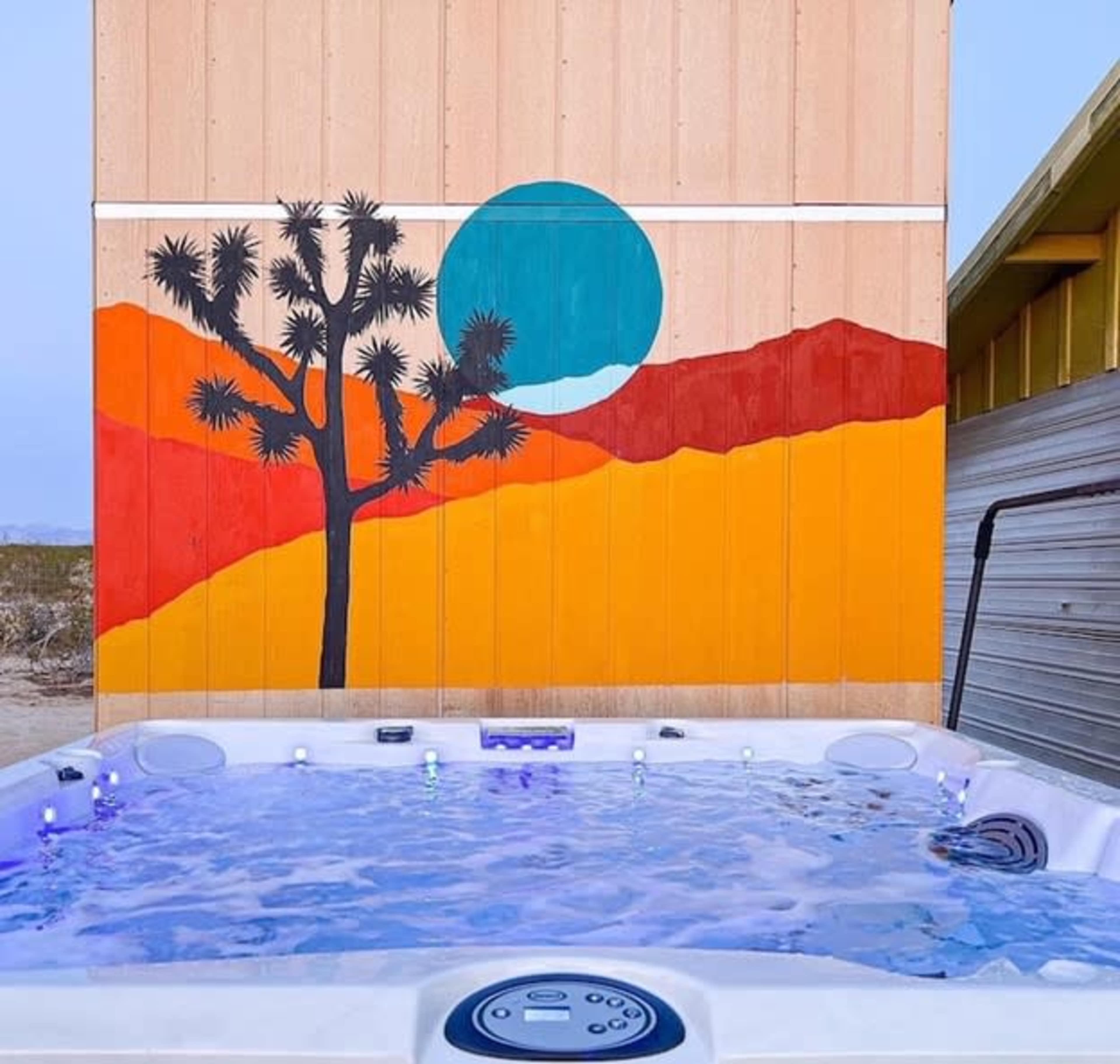 A hot tub in front of a colorful mural depicting a tree and a sunset in a desert landscape.