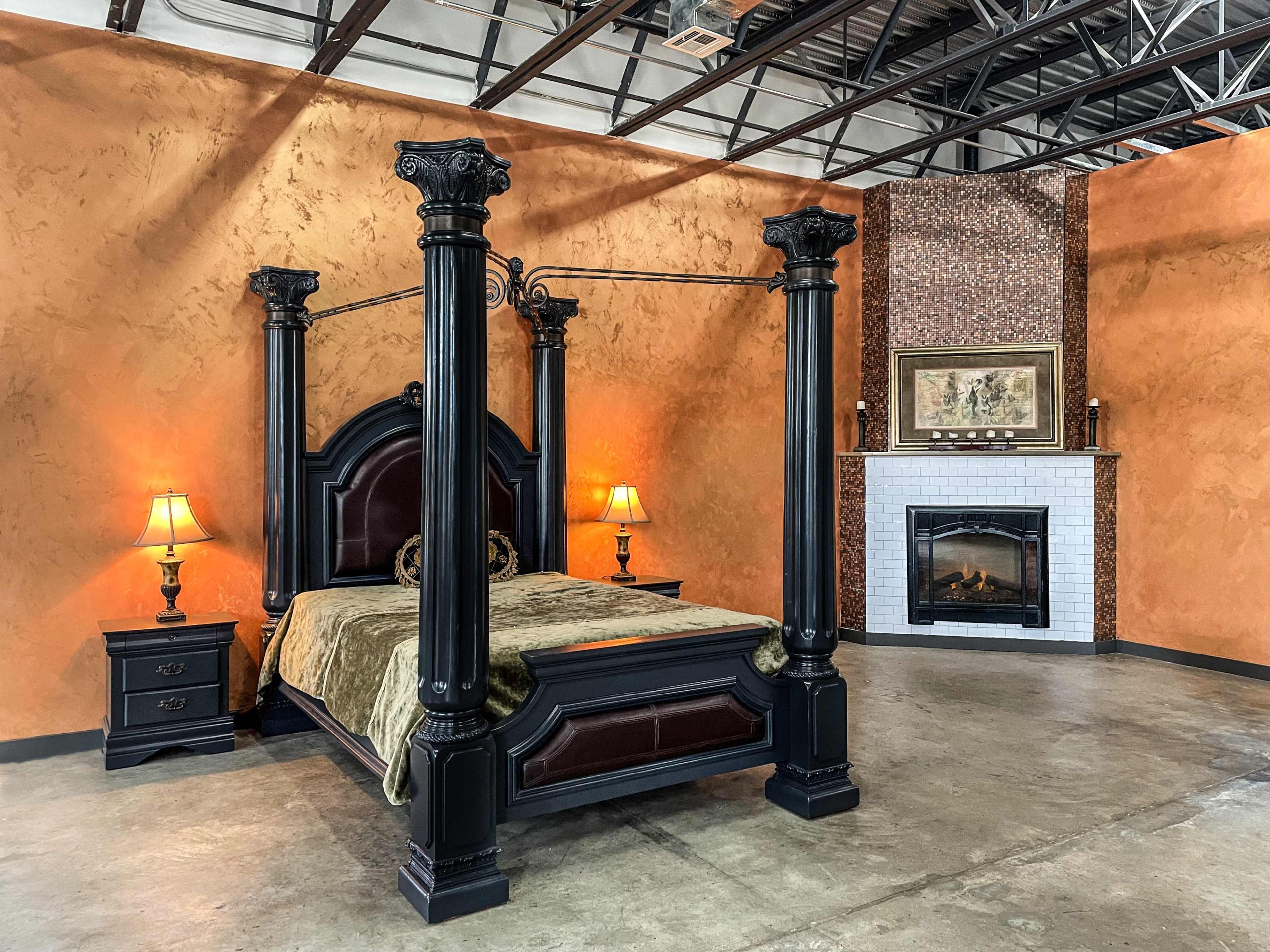 A four-poster bed with black wooden posts, flanked by two bedside lamps, set against an orange wall with a fireplace and artwork in the background.