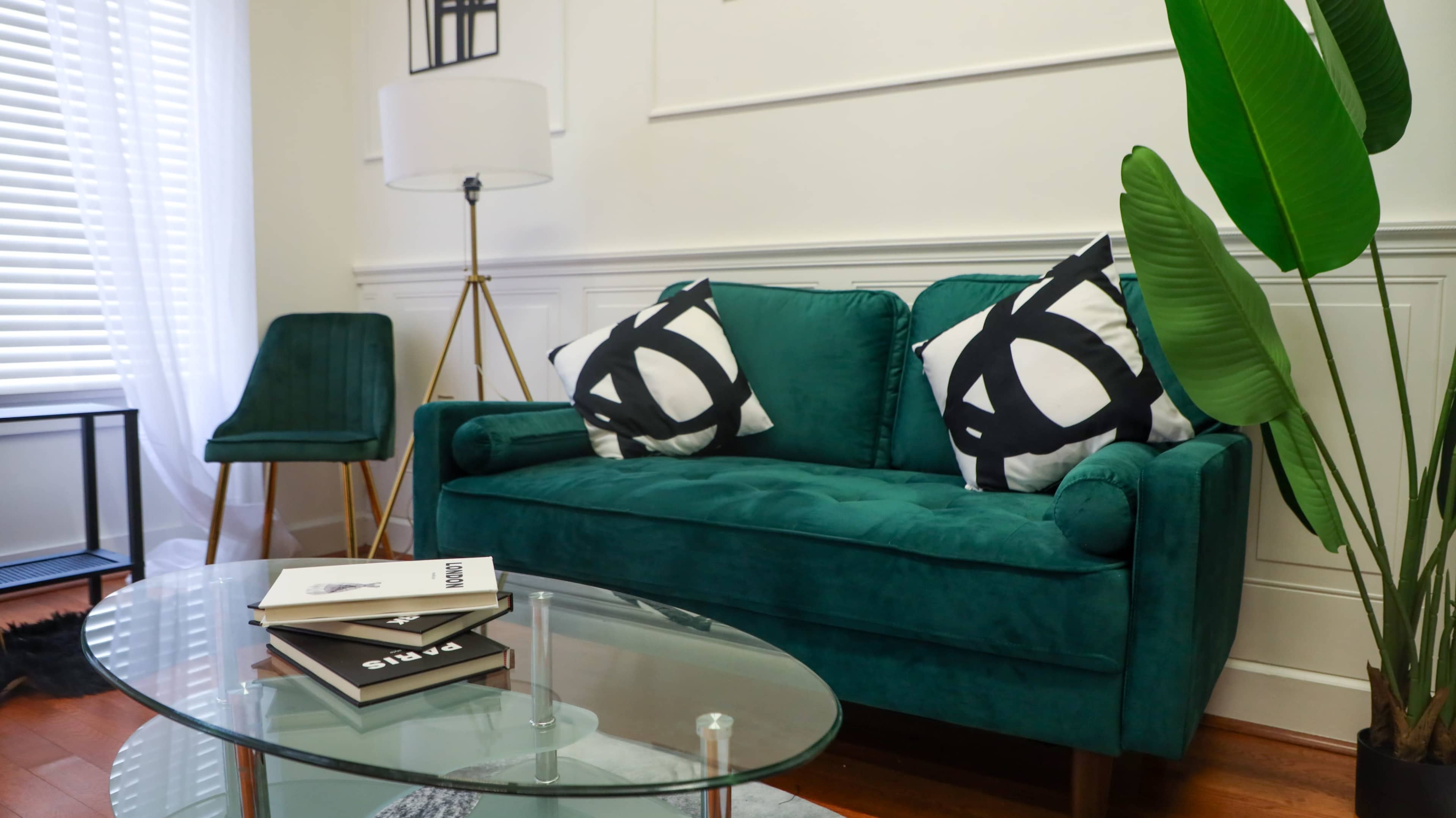 A green velvet sofa with decorative black and white pillows is positioned near a glass coffee table and a lamp, with a potted plant and a chair in the background.