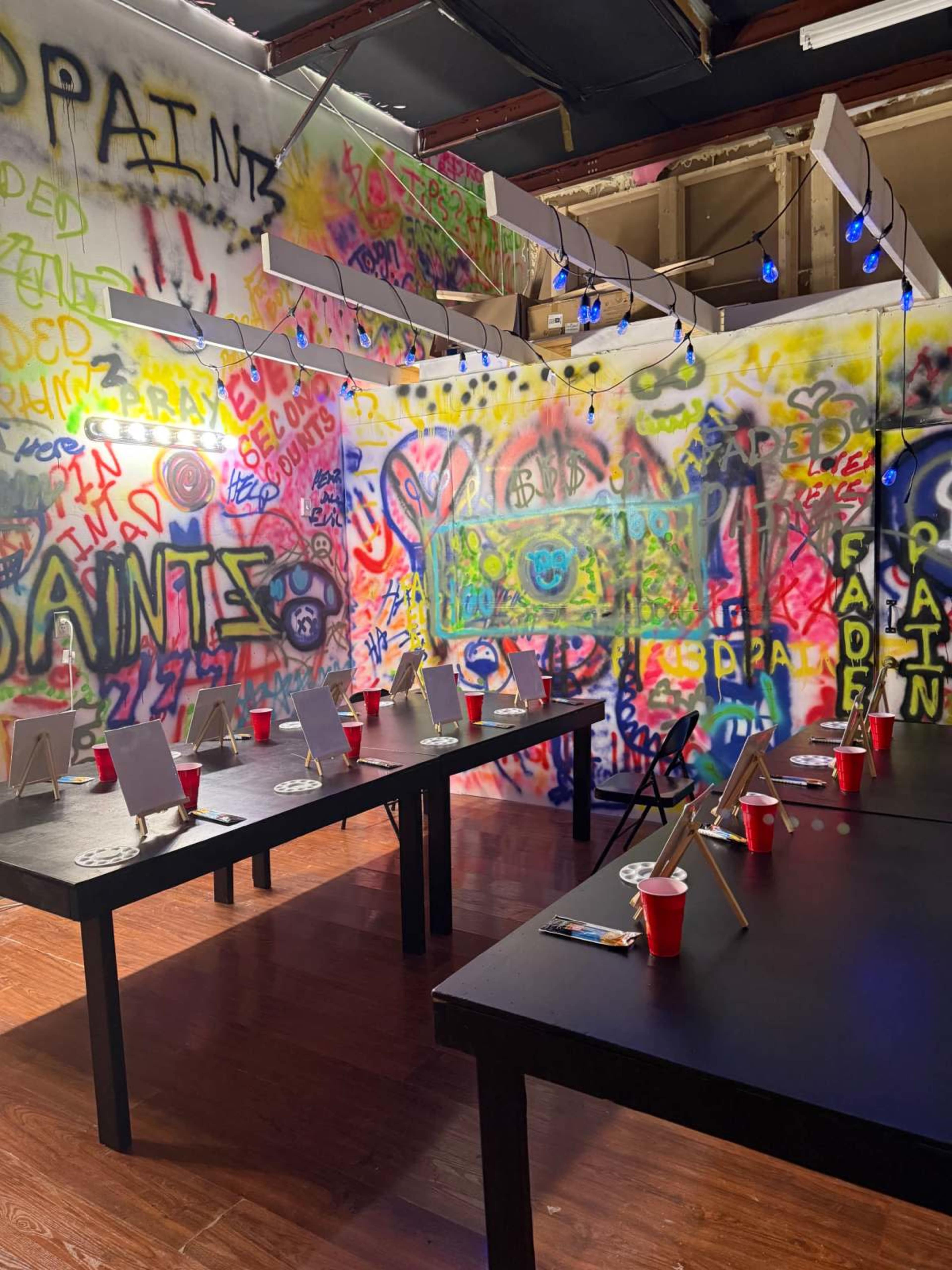 The image shows an indoor space filled with colorful graffiti on the walls, with several black tables set up, each holding red cups and blank canvases.
