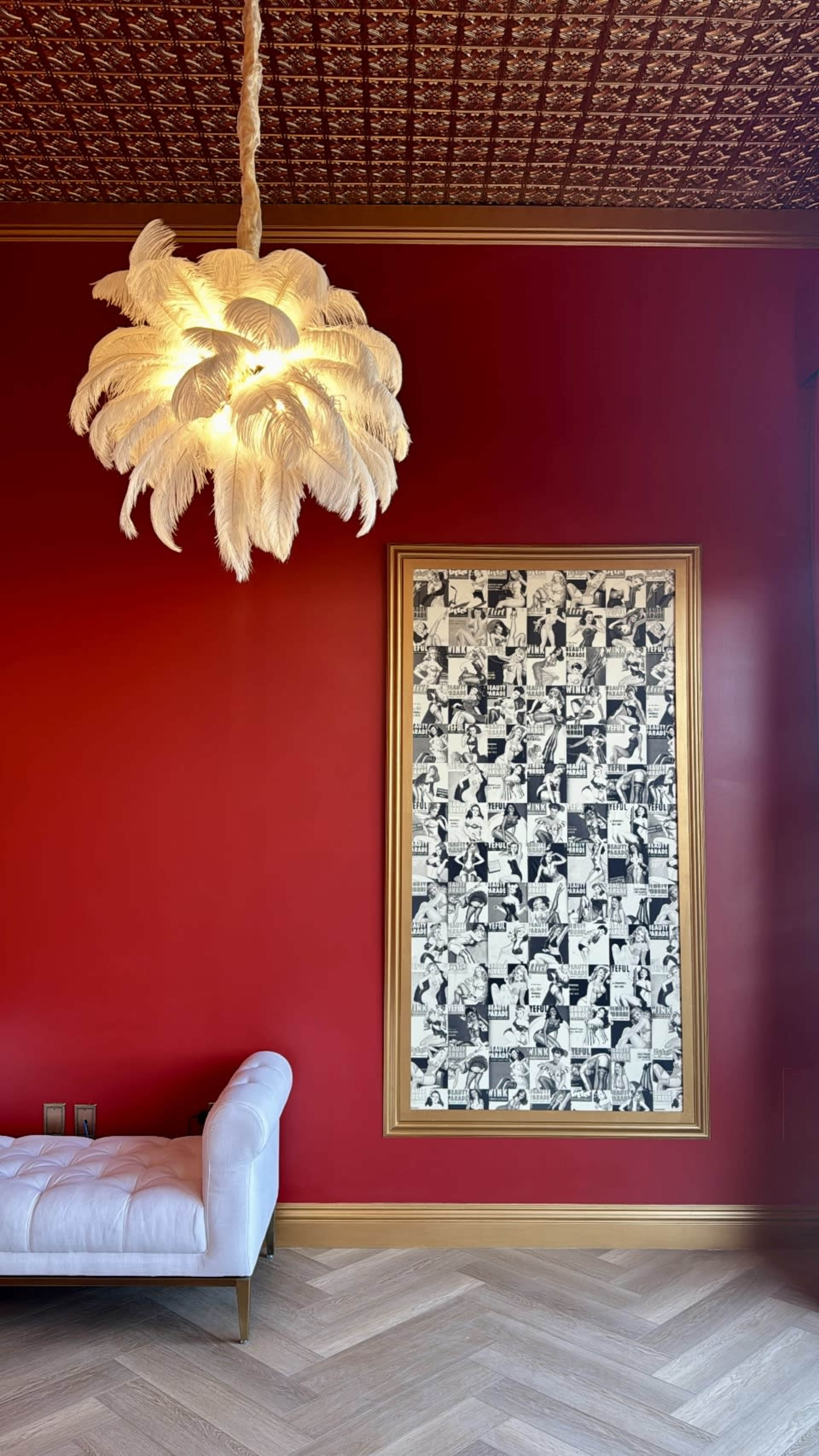 A white, feather-like chandelier hangs from a patterned ceiling above a white chaise lounge, next to a large, framed black-and-white artwork on a red wall.