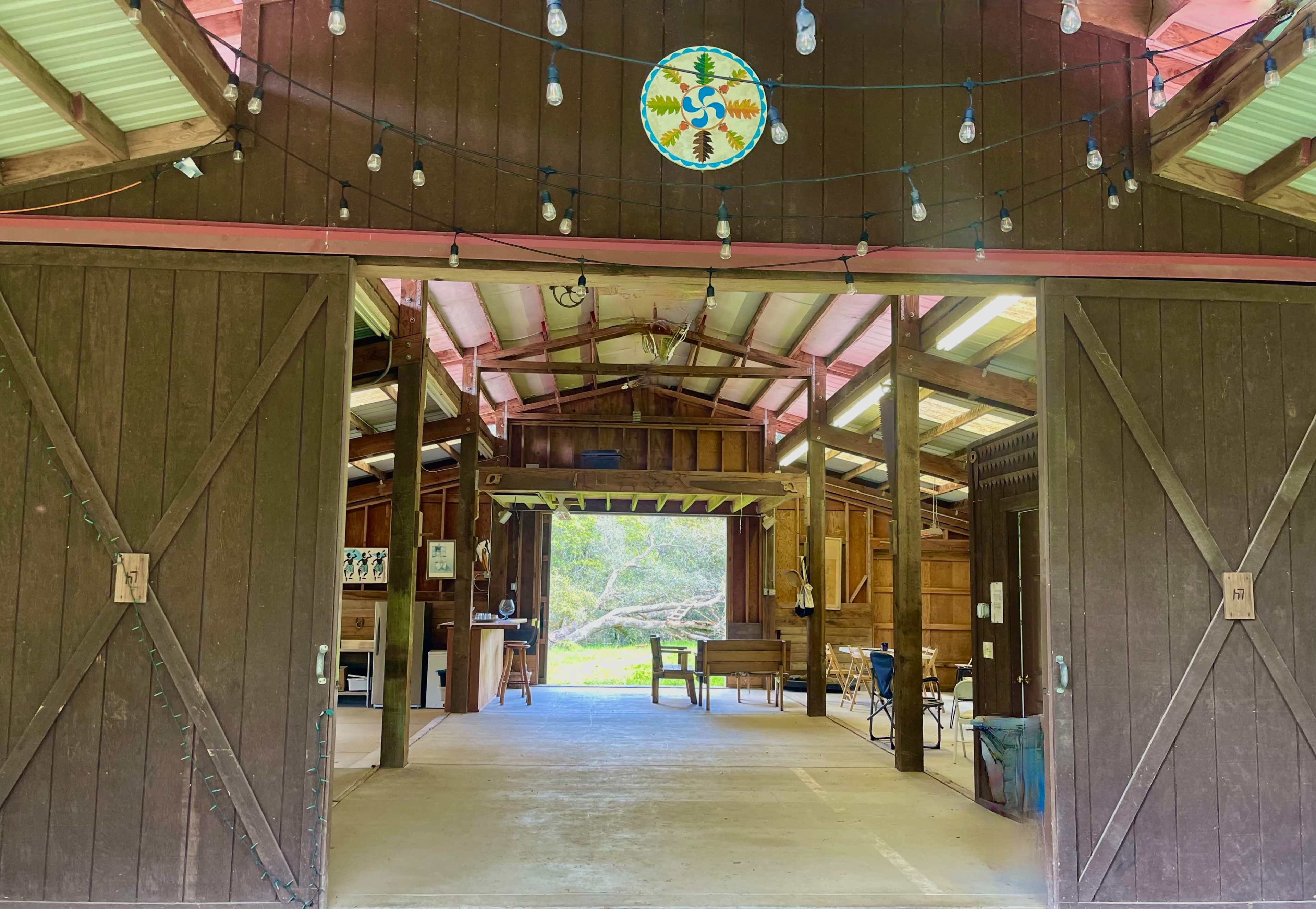 The image shows an open barn structure with wooden walls and string lights hanging from the ceiling, revealing tables and chairs inside.