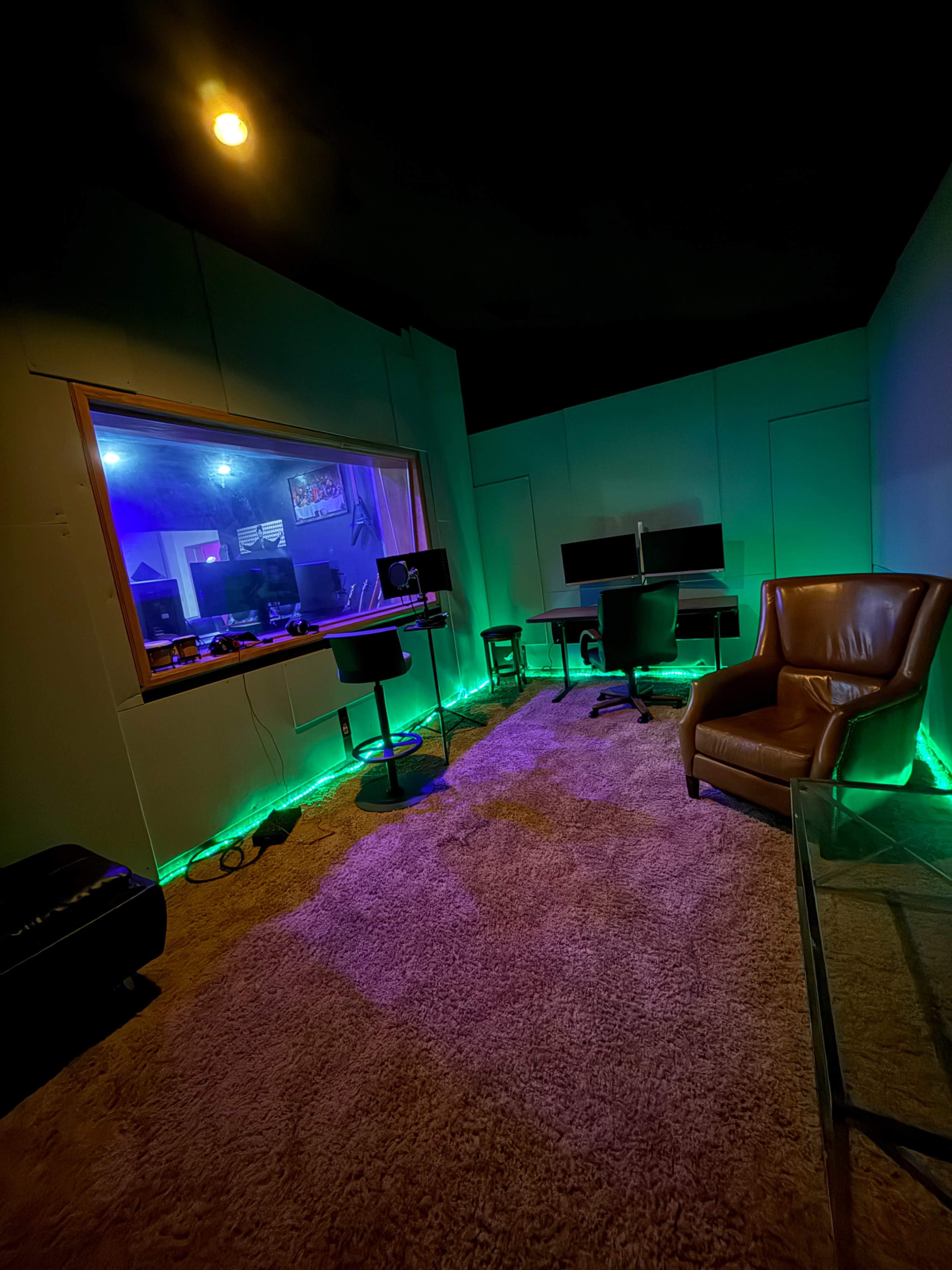 A modern recording studio features a cozy brown leather chair and a glass table, illuminated by colorful LED lights against the walls.