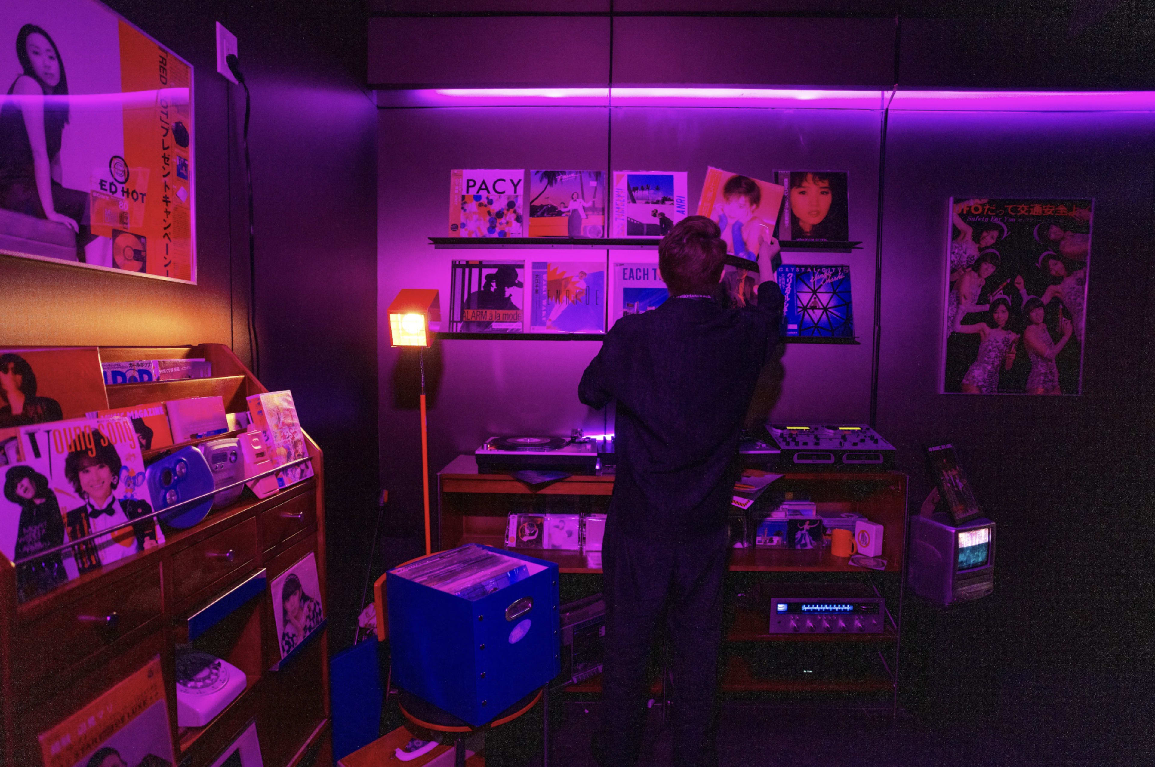 A person stands in a dimly lit room filled with vinyl records and colorful ambient lighting, adjusting a turntable.