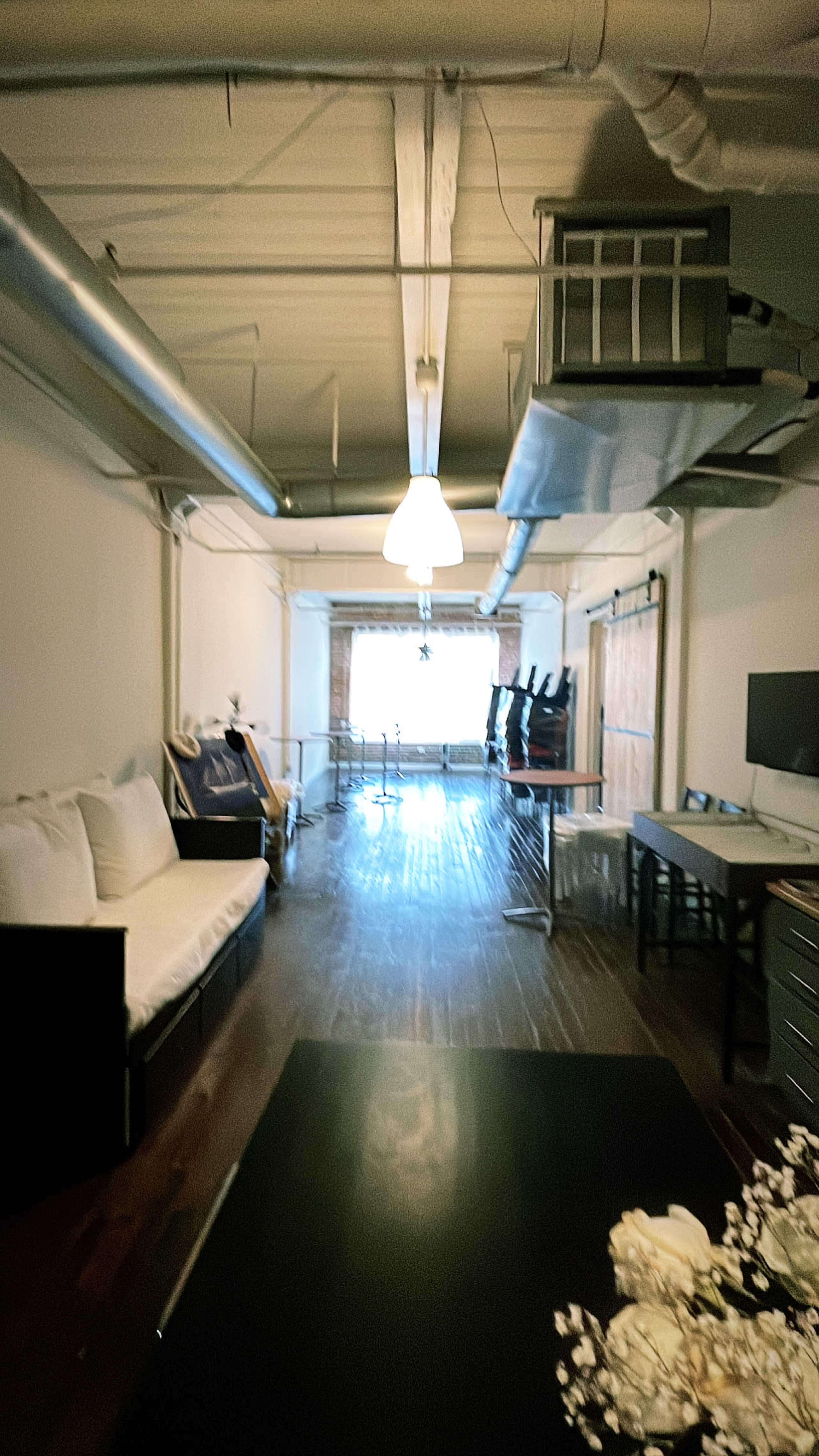 A long, narrow room with wooden floors, a sofa on one side, and desks along the other, leading to a window at the end.