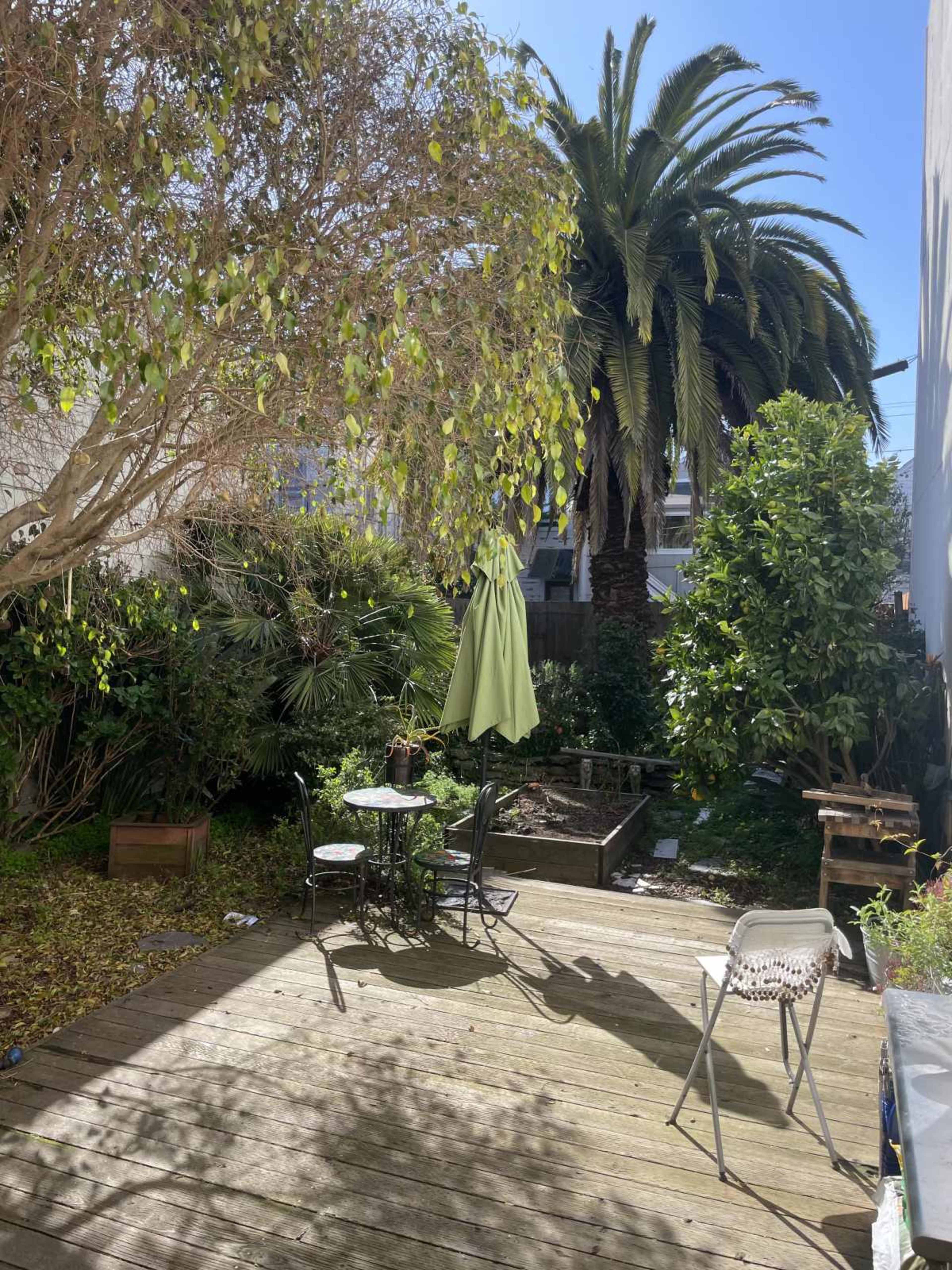 The image shows a backyard garden with wooden decking, a small circular table and chairs, a green umbrella, and palm trees surrounded by various plants.