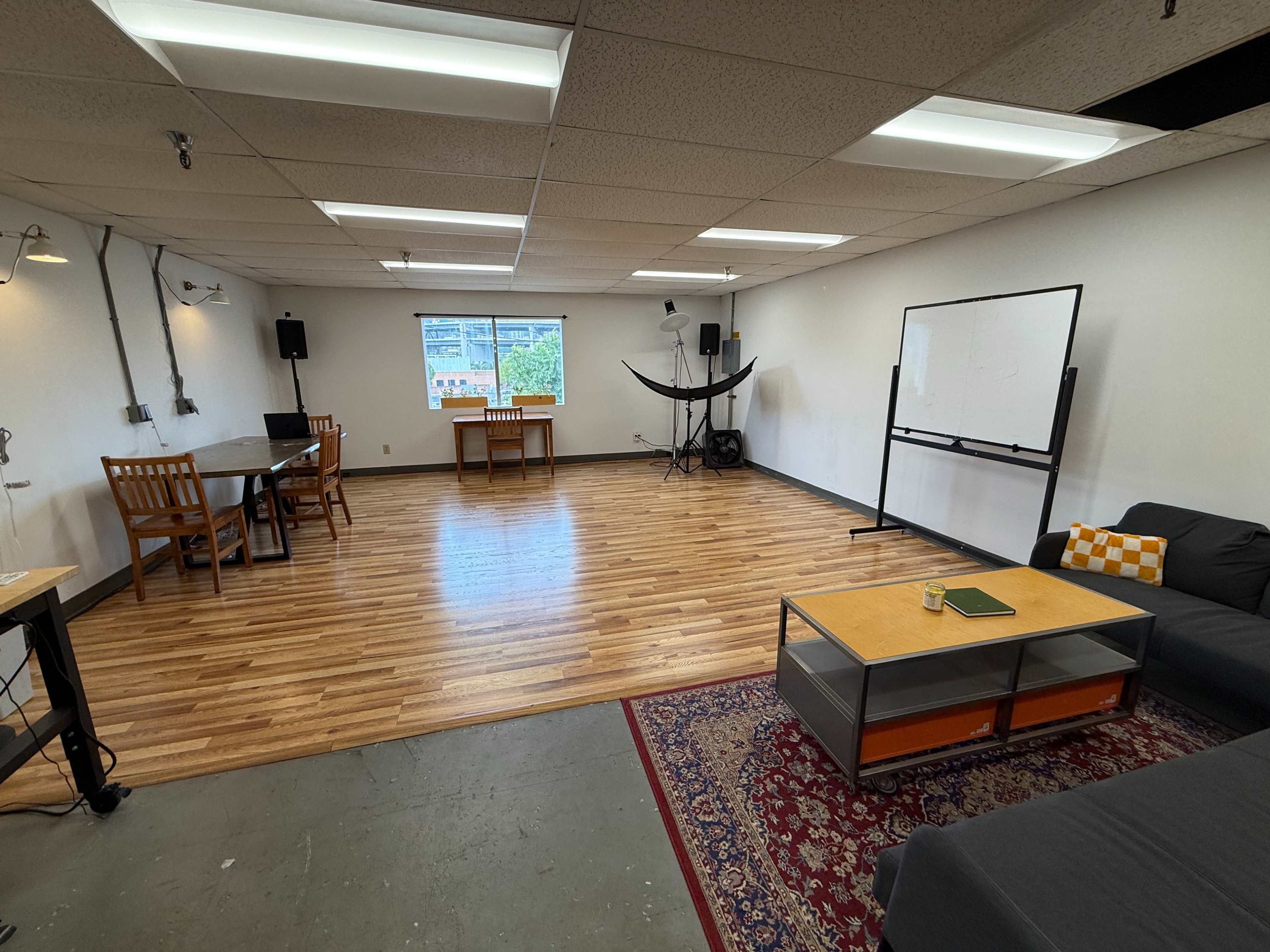 The image shows an open, well-lit room with hardwood flooring, featuring a couch, a coffee table, a whiteboard, and several tables and chairs arranged throughout the space.