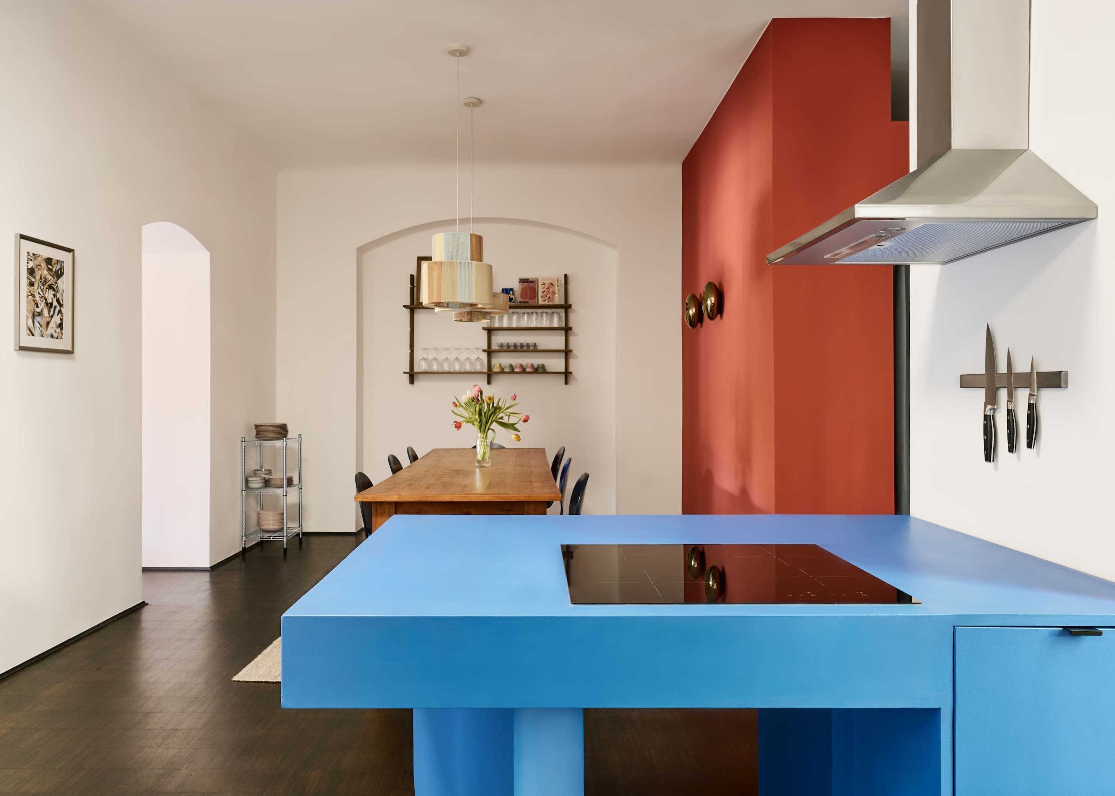 The image shows a modern kitchen featuring a blue island with a cooktop, a wooden dining table in the background, and a red accent wall.