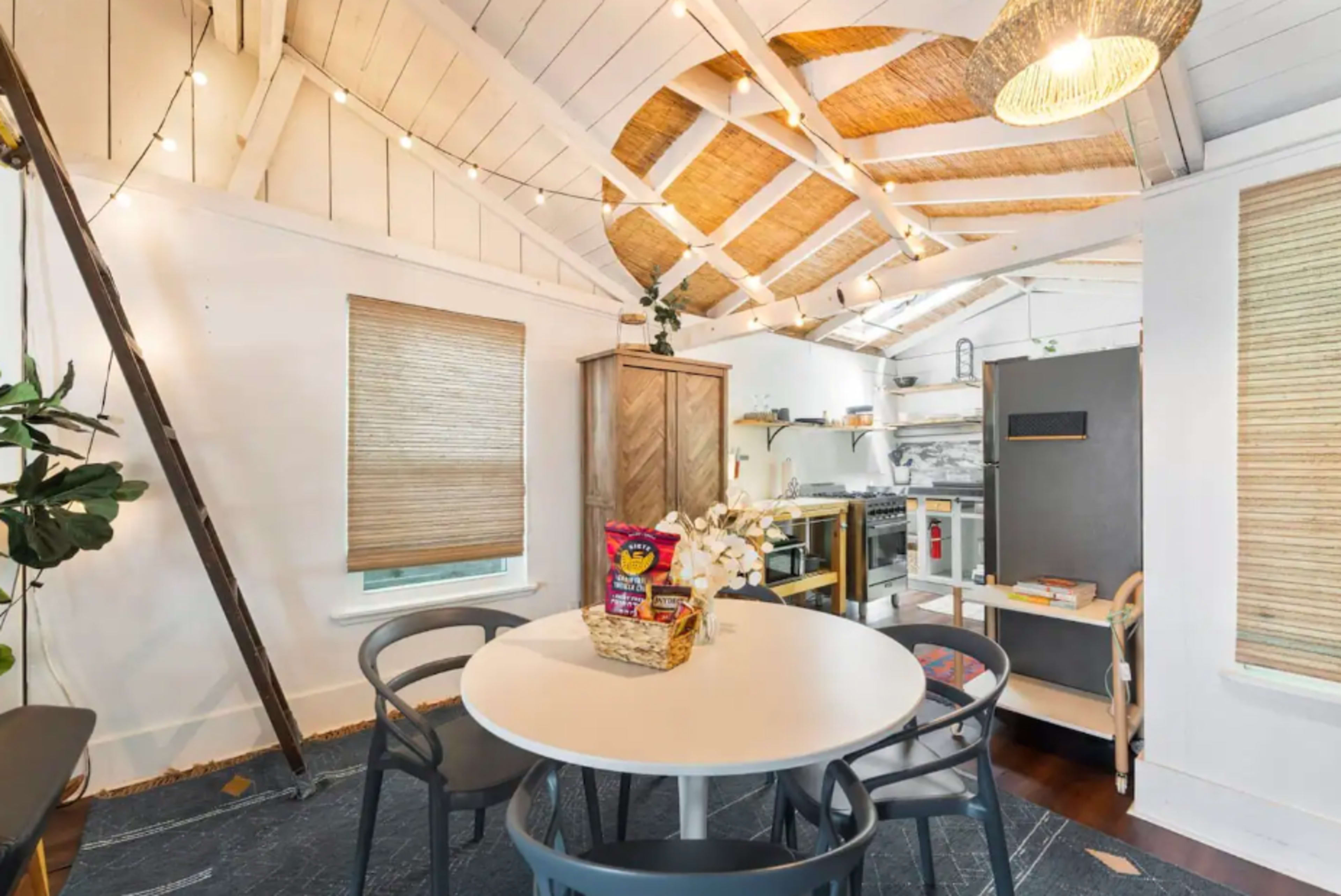 A bright kitchen and dining area featuring a round white table surrounded by gray chairs, with a wooden ceiling and various kitchen appliances in the background.