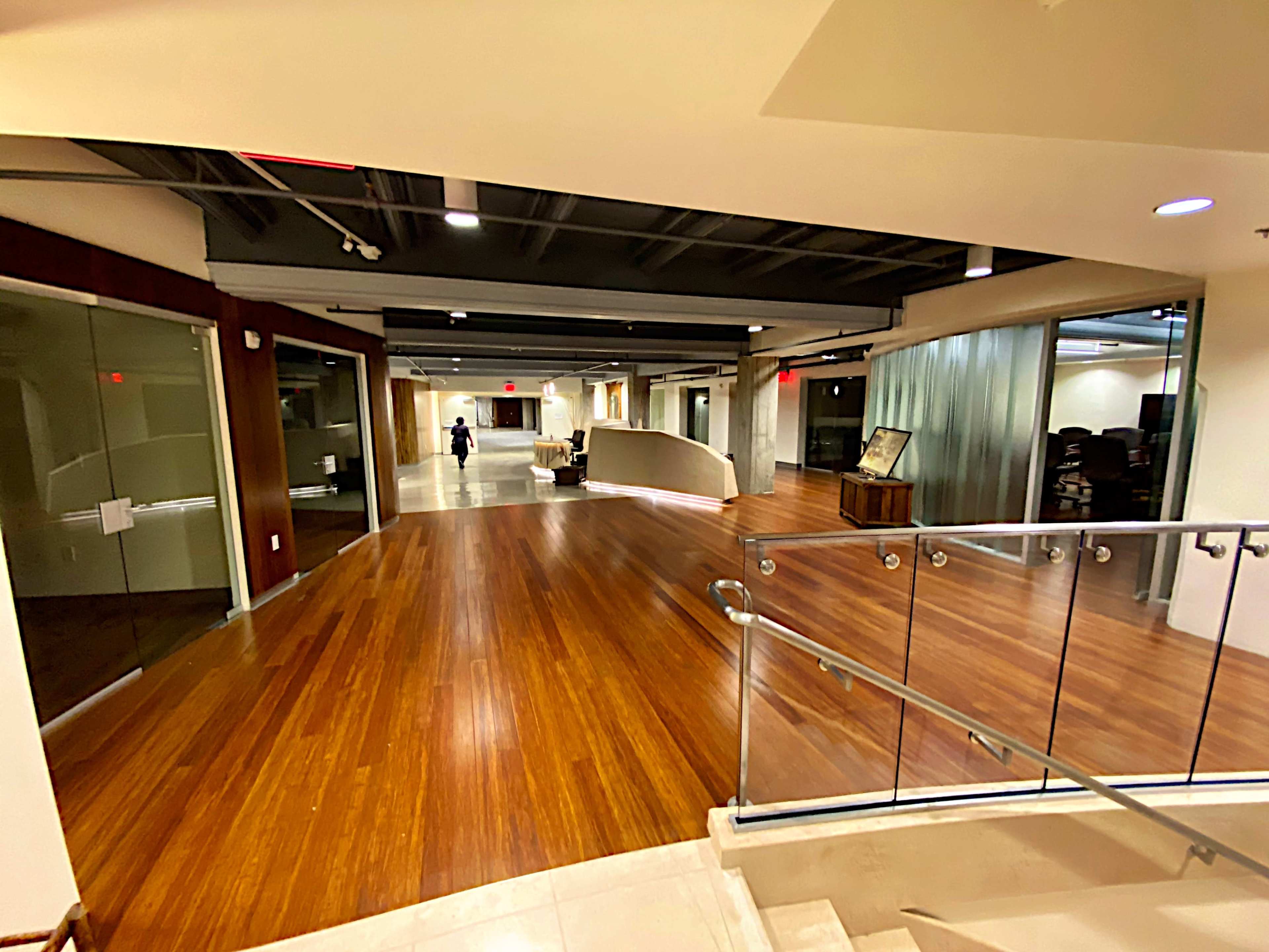 The image shows a modern office corridor with polished wooden floors and glass-walled meeting rooms.