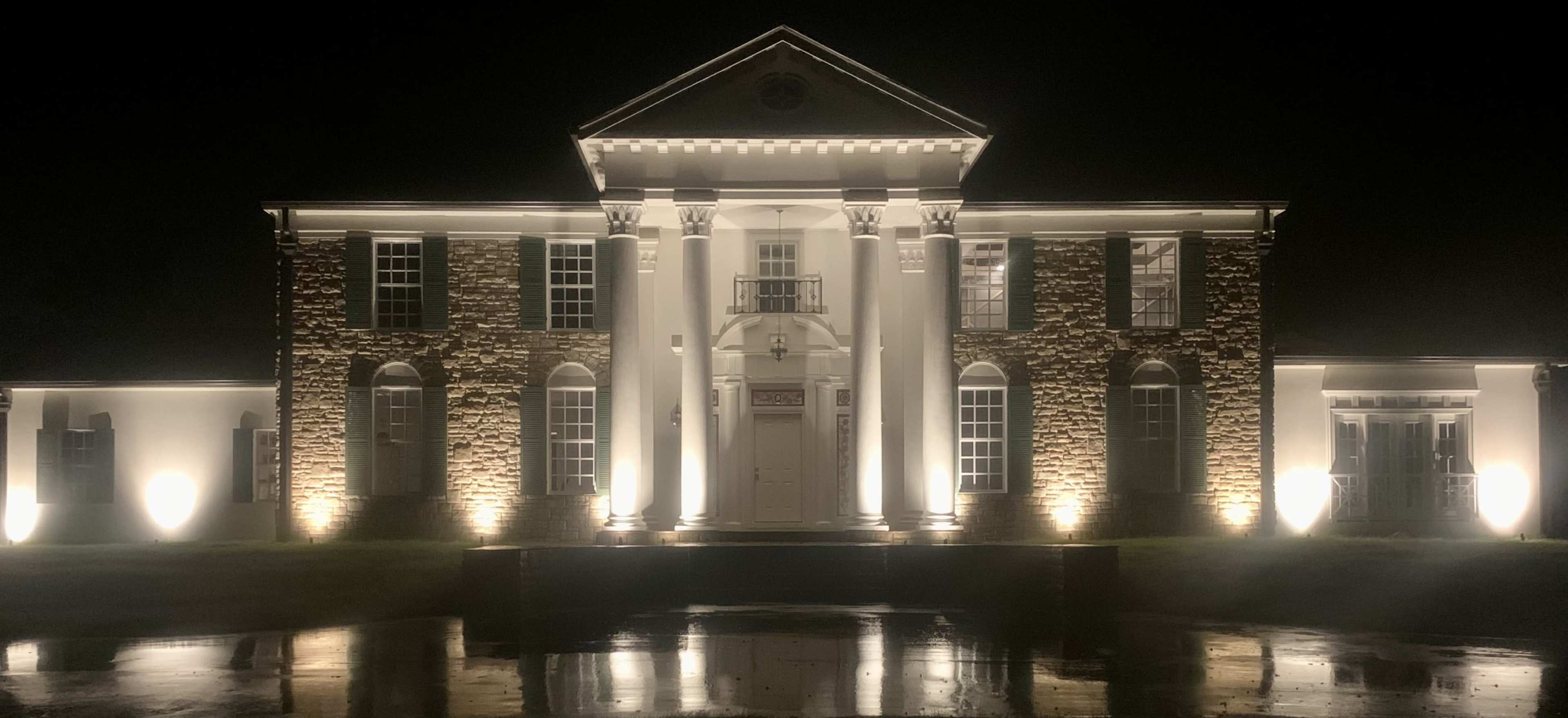 A large, stately mansion is illuminated at night, with prominent architectural features and a reflecting pond in front.