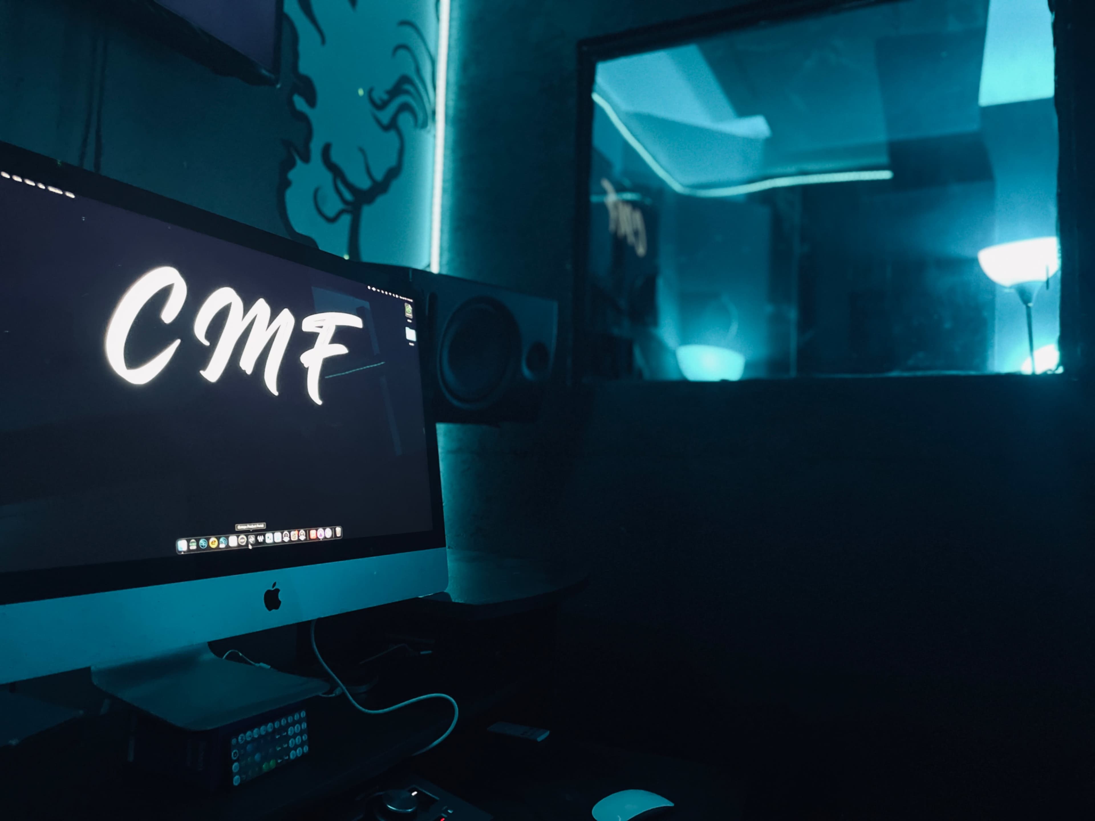 A dimly lit recording studio features an iMac displaying "CMF," with blue ambient lighting and a glass window to a soundproof booth.