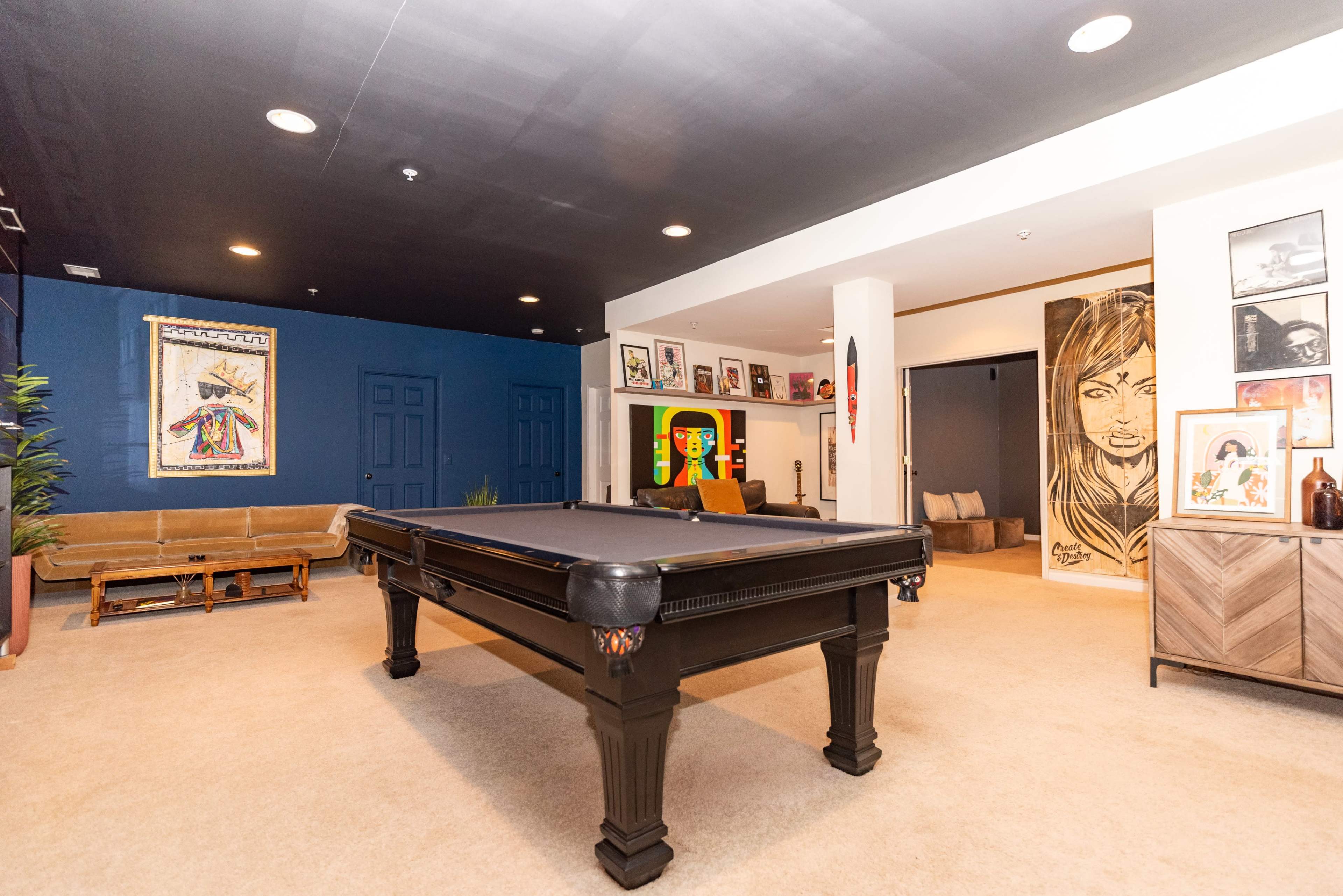 The image shows a spacious room featuring a black pool table at the center, surrounded by various artworks and seating areas.