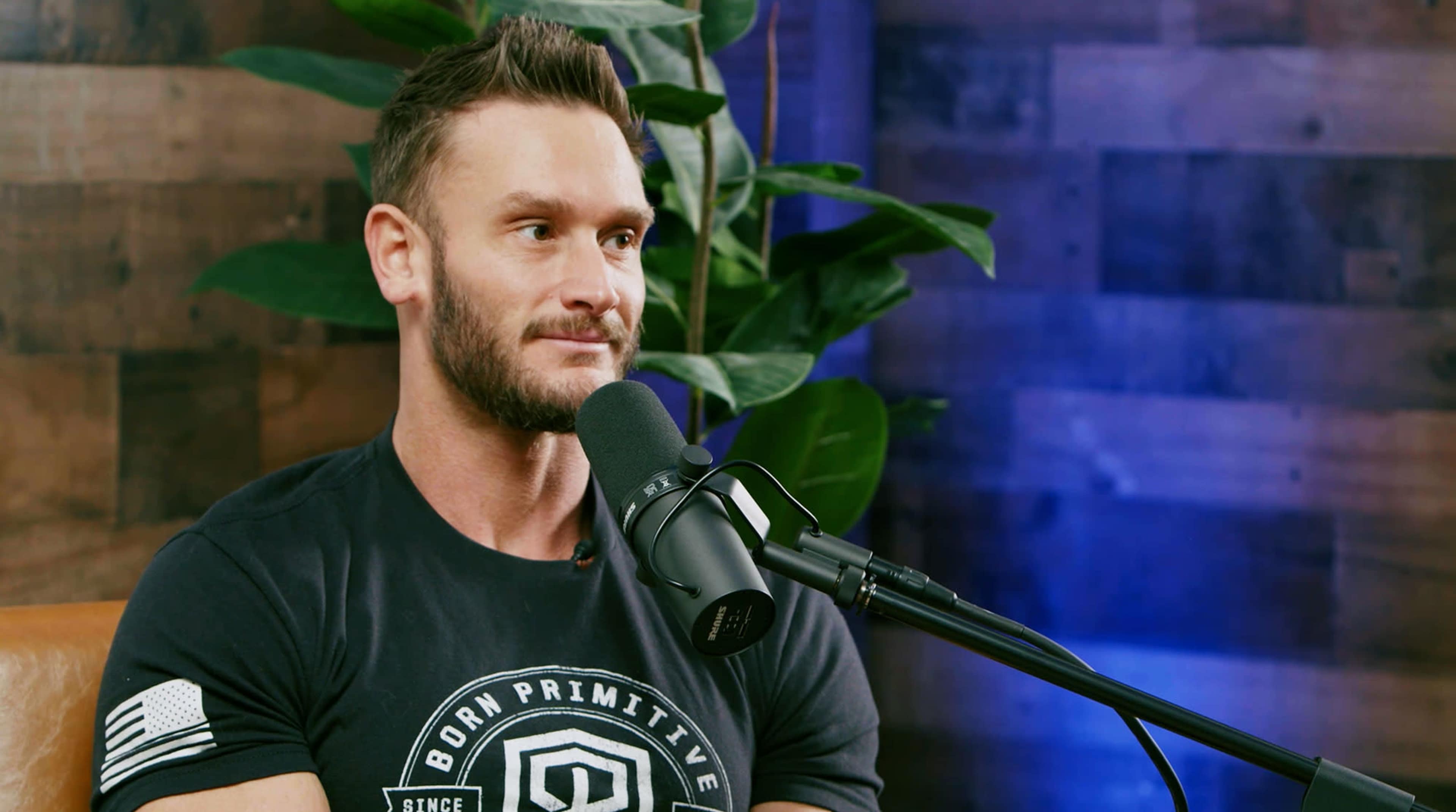A man with a beard sits in front of a microphone, surrounded by greenery and wooden decor.