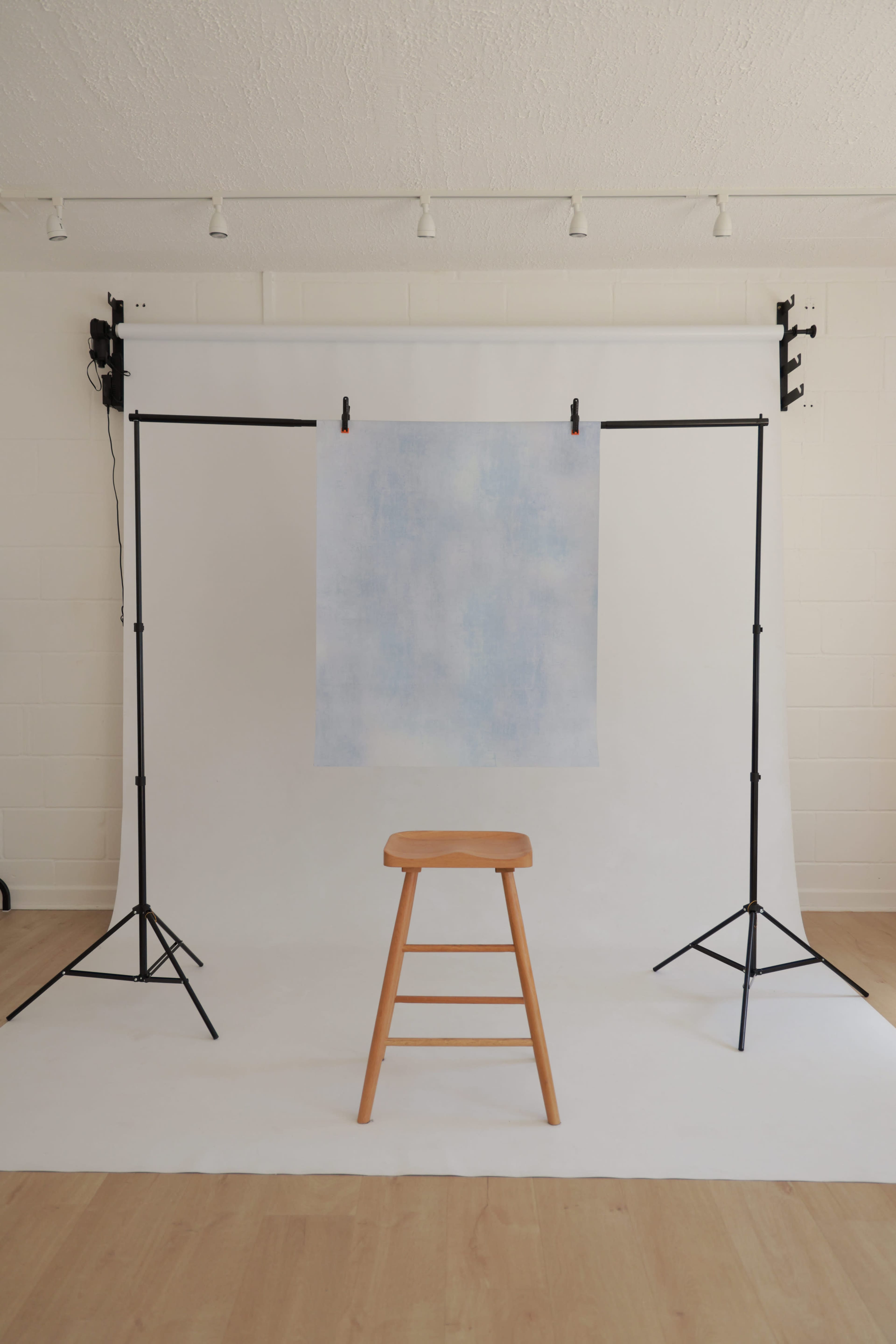 A wooden stool is positioned in front of a light blue backdrop set up on a stand in a simple, well-lit studio space.