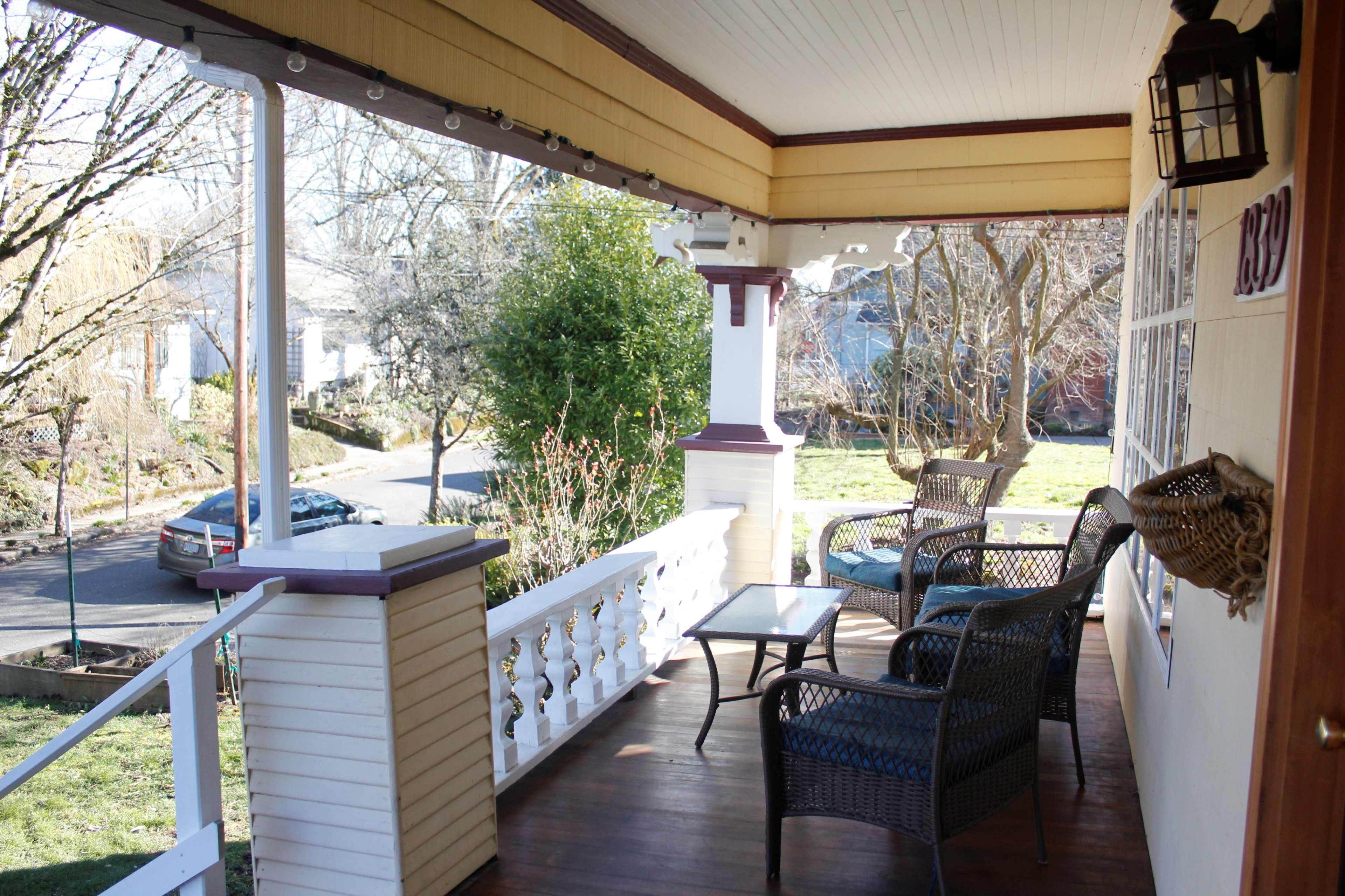 A covered porch with wicker chairs and a small table, overlooking a quiet street lined with trees.