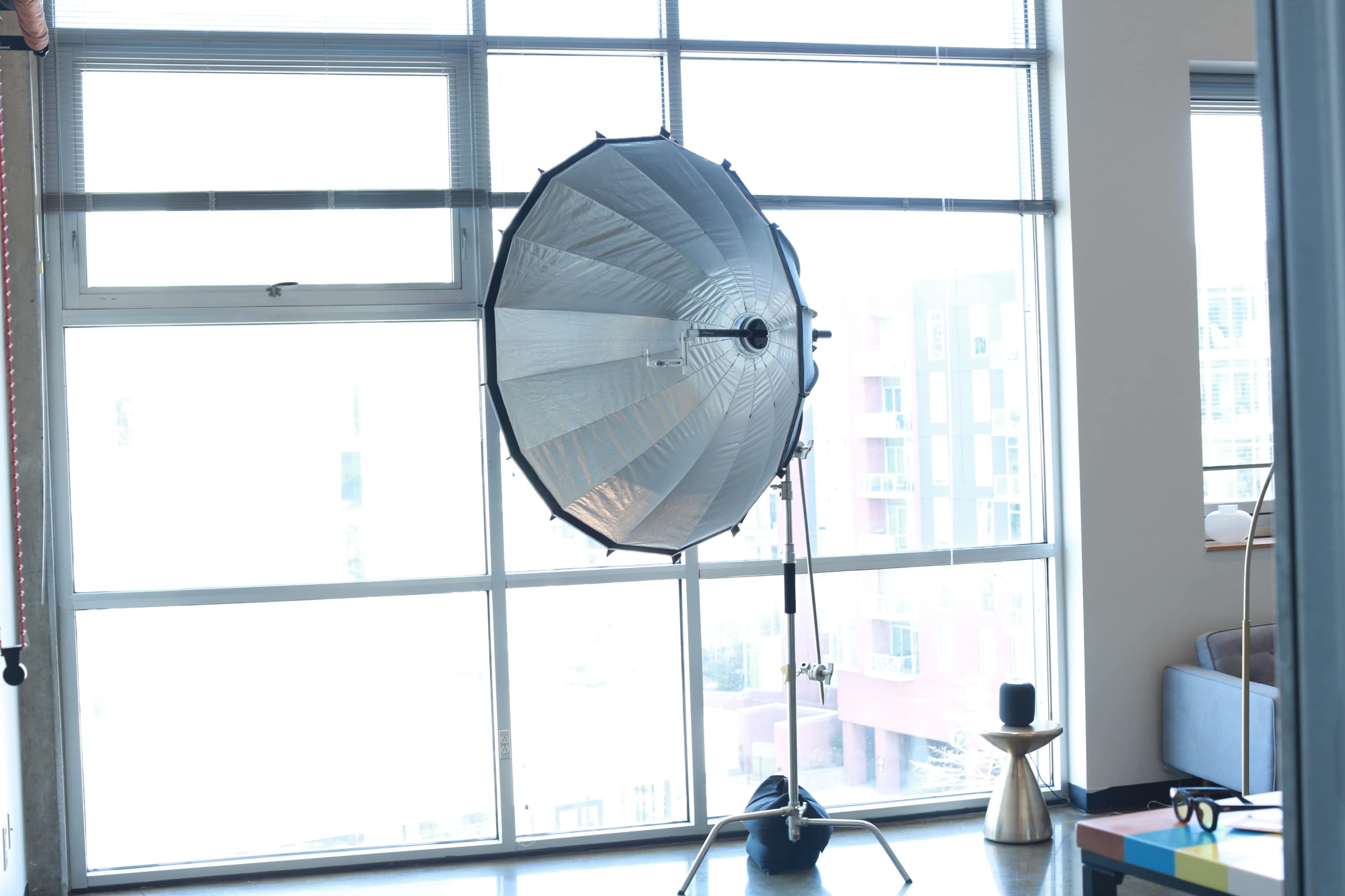 A large silver umbrella softbox is set up in front of a window in a bright room.