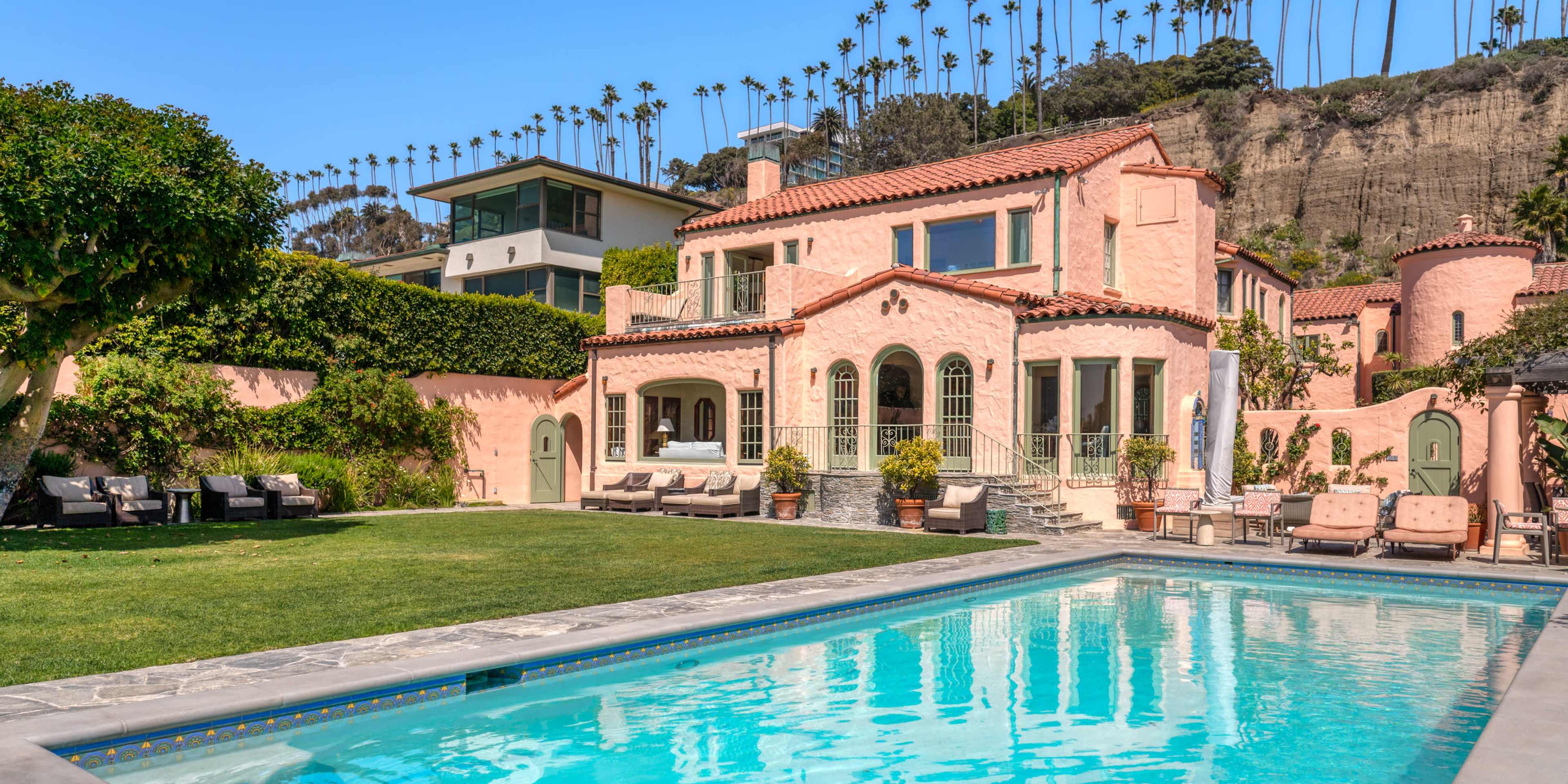 A two-story pink stucco house with arched windows and a pool is surrounded by a manicured lawn and palm trees.