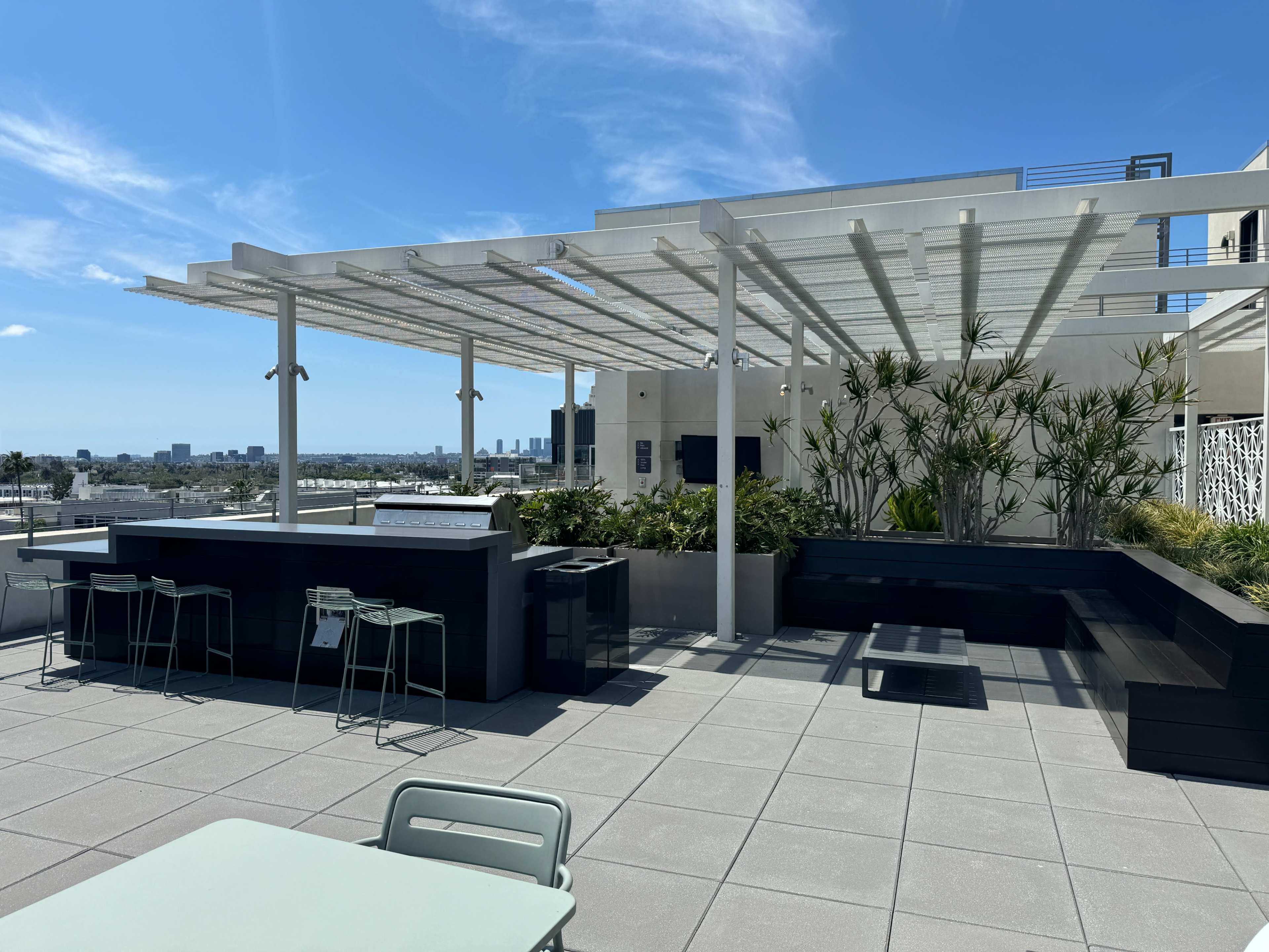 A rooftop terrace featuring a bar area, seating, and potted greenery, with a city skyline visible in the background.