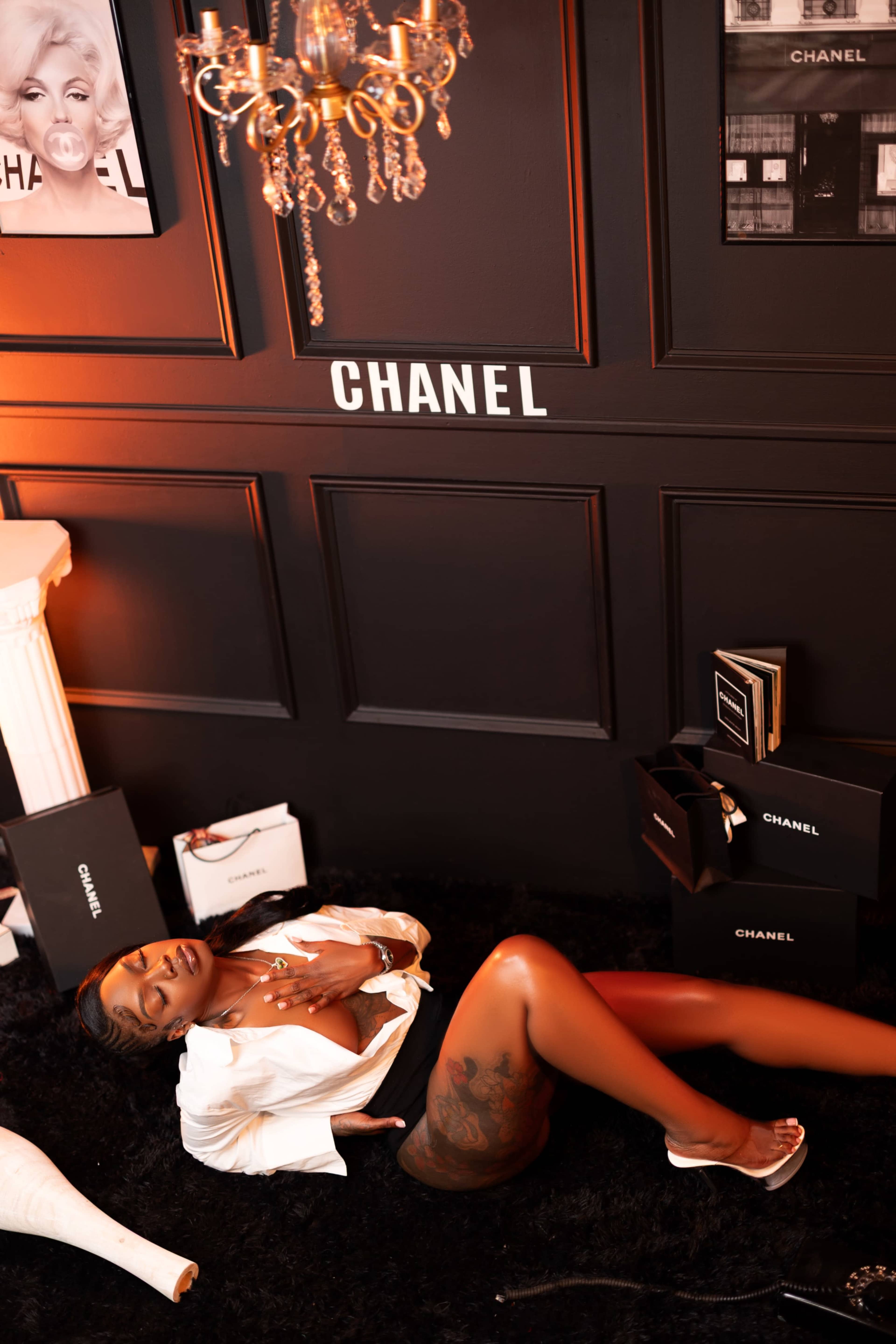 A person lies on a black carpet surrounded by Chanel boxes and a chandelier, with a decorative wall in the background.