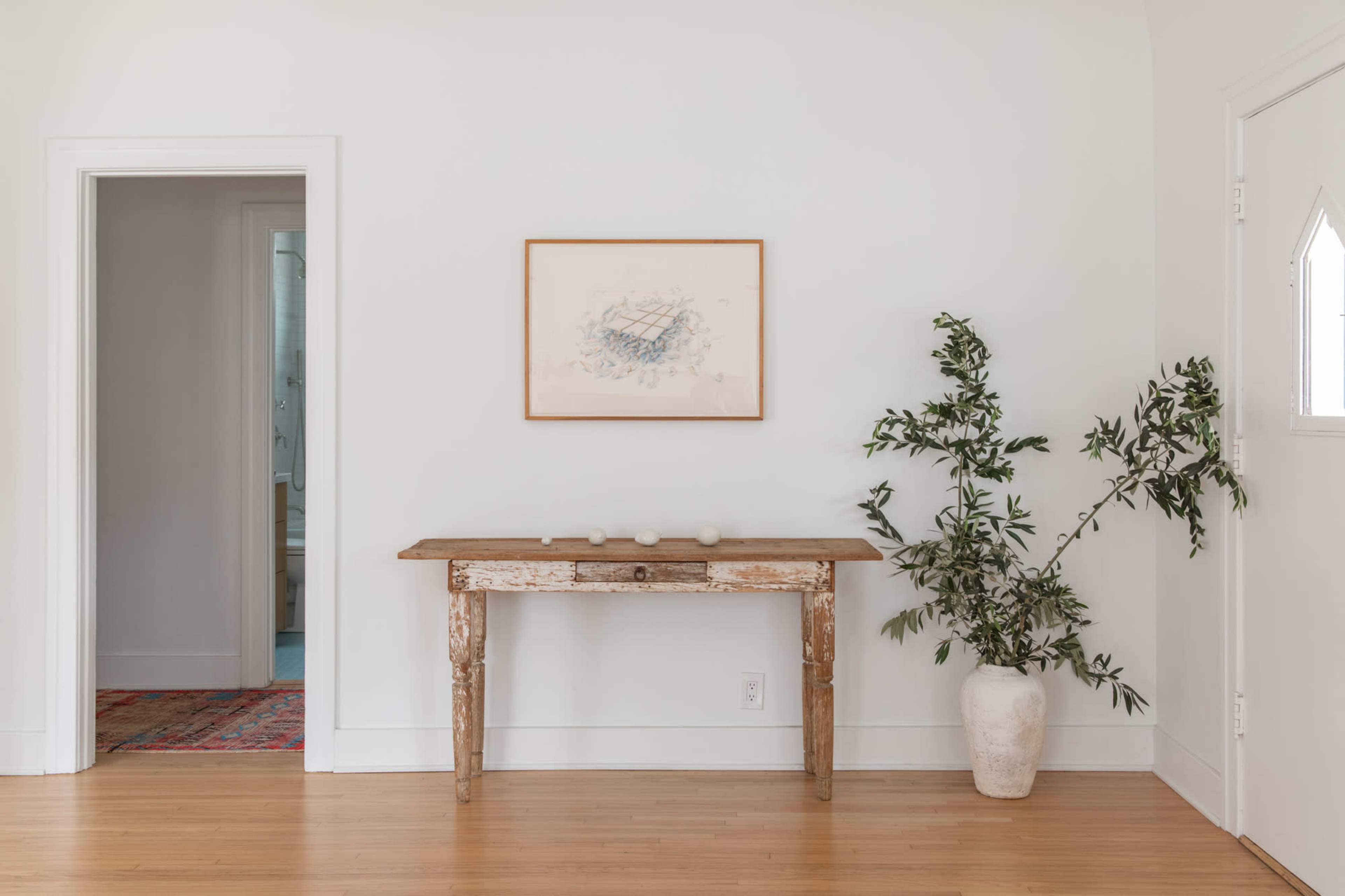 A bright hallway featuring a wooden table with three small decorative items, a framed artwork on the wall, and a potted plant beside it.
