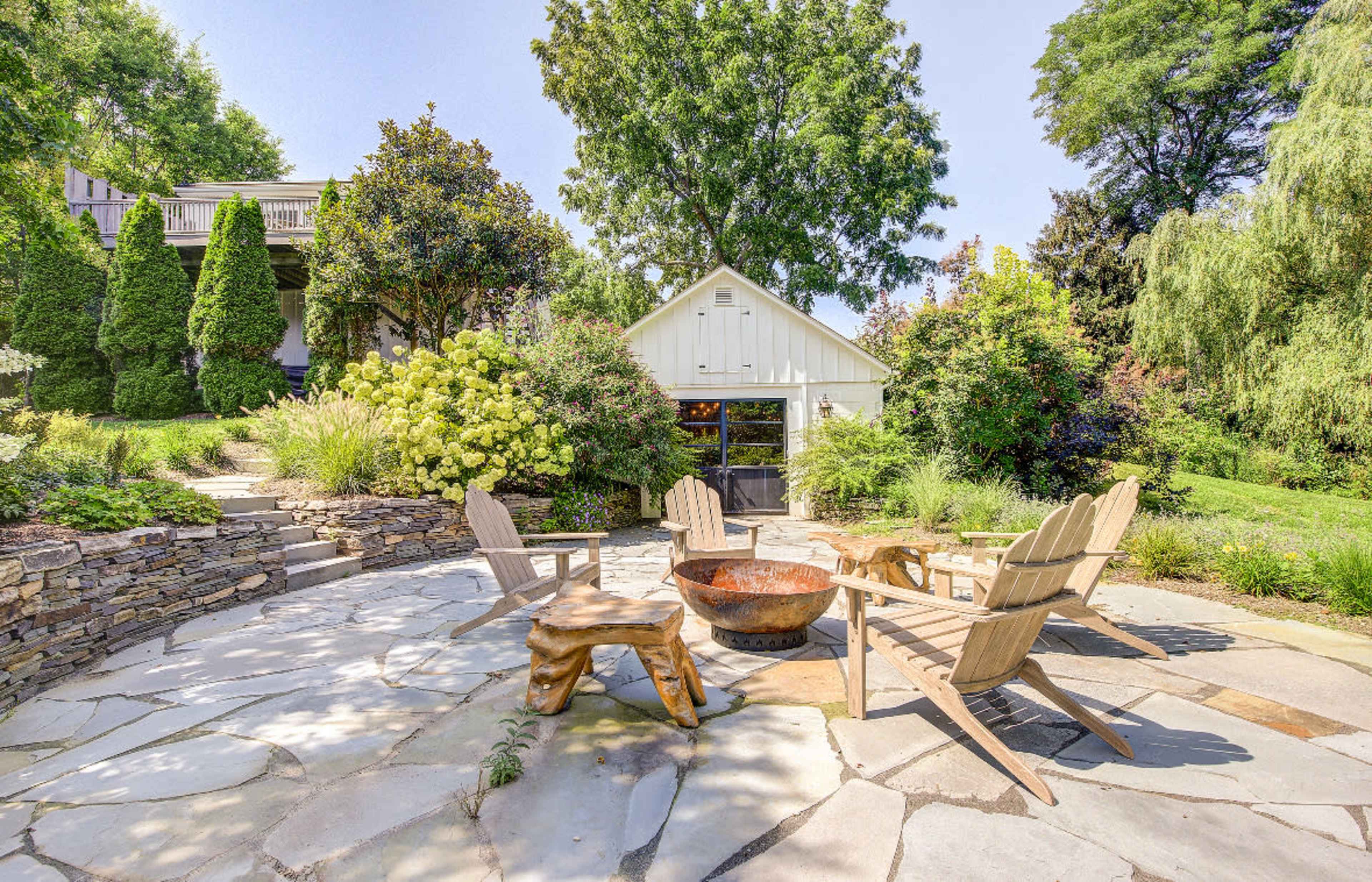 The image shows a stone patio with wooden adirondack chairs arranged around a fire pit, surrounded by lush greenery and a white shed in the background.