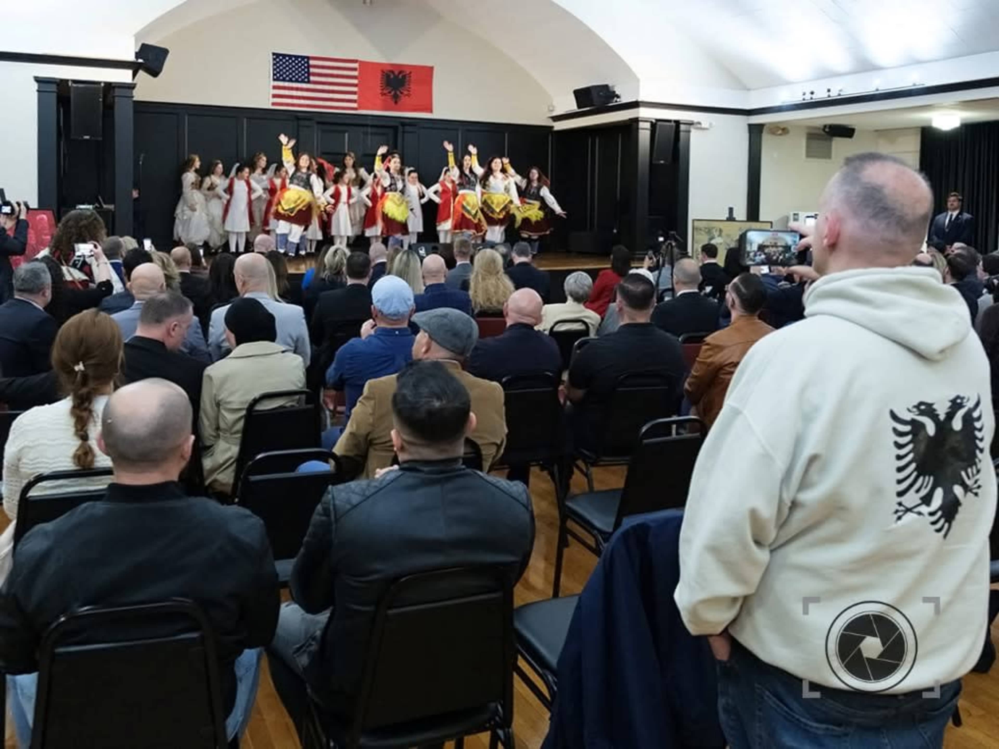 A group of performers dressed in traditional clothing is dancing on stage in front of an audience seated in a hall decorated with American and Albanian flags.