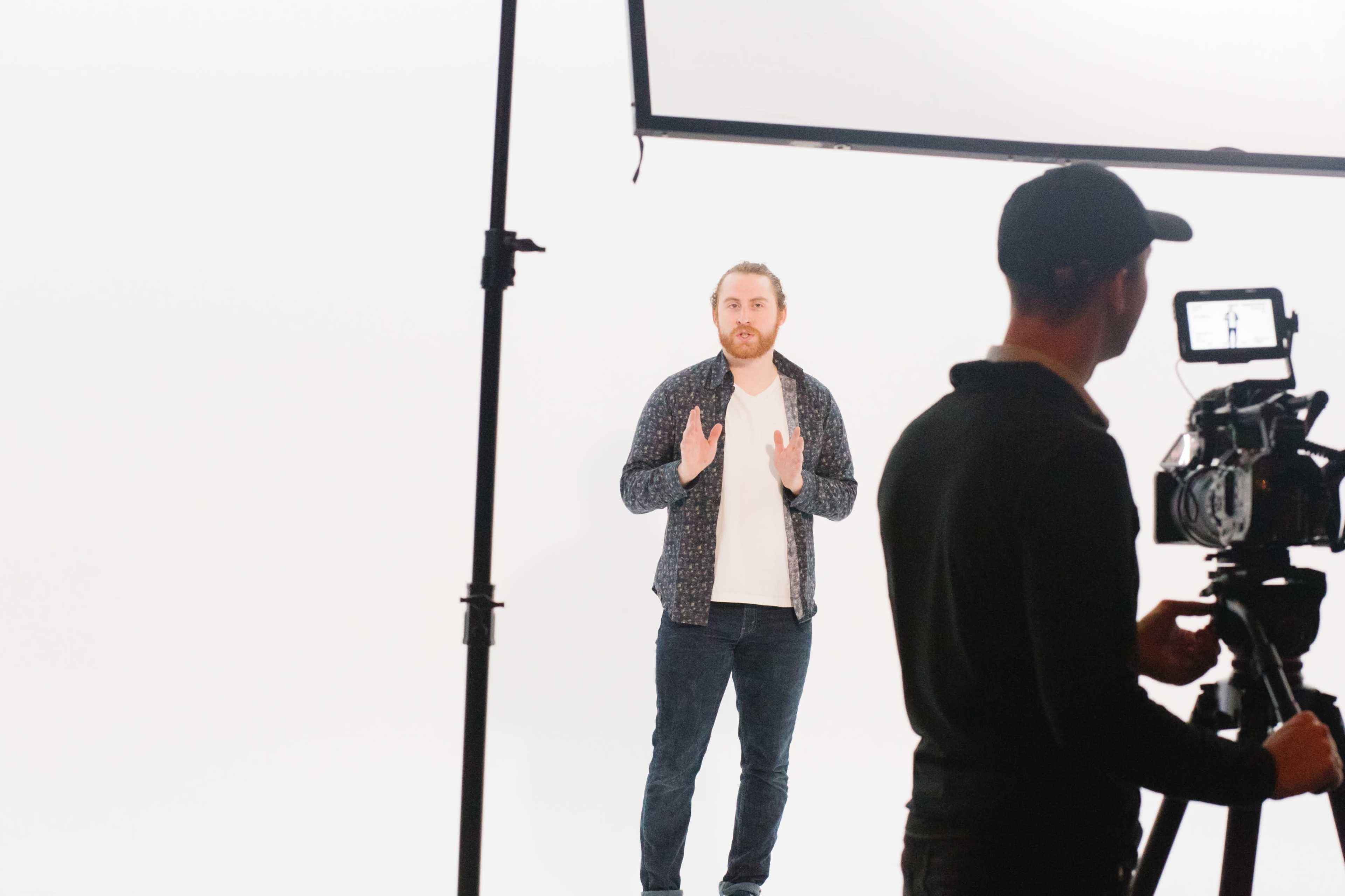 A man is speaking in front of a white backdrop while another person operates a camera on a tripod.