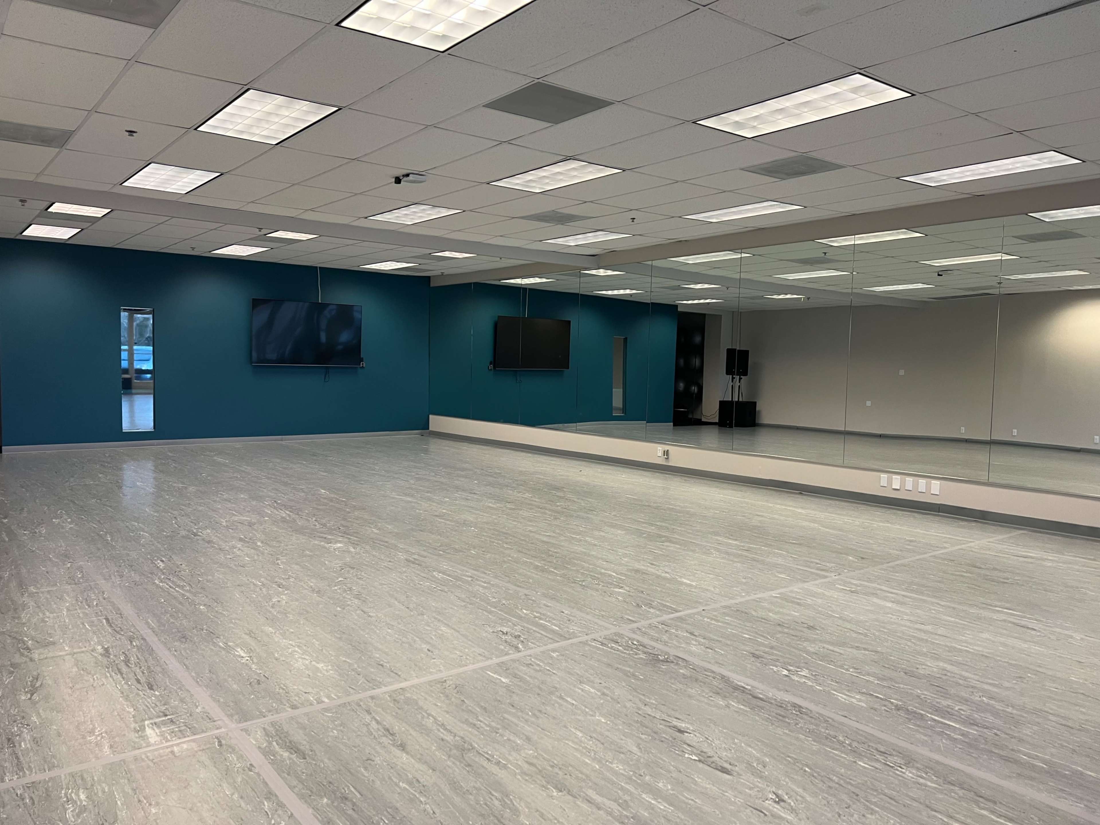 An empty dance studio with large mirrors, two wall-mounted screens, and a light-colored floor.